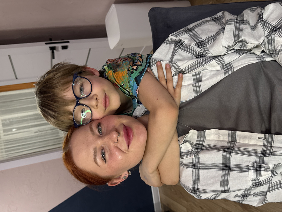 The image displays an adult female and a male child in an interior residential setting within Rostov-on-Don, Russia. The female, seated, possesses reddish-brown hair and a septum piercing, wearing a light-colored plaid shirt over a dark grey t-shirt. She has visible makeup, including winged eyeliner and glossy pink lipstick. The male child, approximately 6-10 years old, has light brown hair and wears dark-rimmed glasses and a colorful, patterned long-sleeved shirt. He stands behind the female, embracing her with his arms wrapped around her neck. Both individuals are looking directly at the camera, exhibiting faint smiles. The background includes a wall with a diagonal paint line, separating a light pink/peach upper section from a dark blue/grey lower section. To the right, a white interior door with horizontal dark accents and a black handle is visible, alongside a tall, white, rectangular electronic device. The back of a dark grey upholstered chair is partially visible on the right, and light-colored wooden flooring is seen in the lower left.