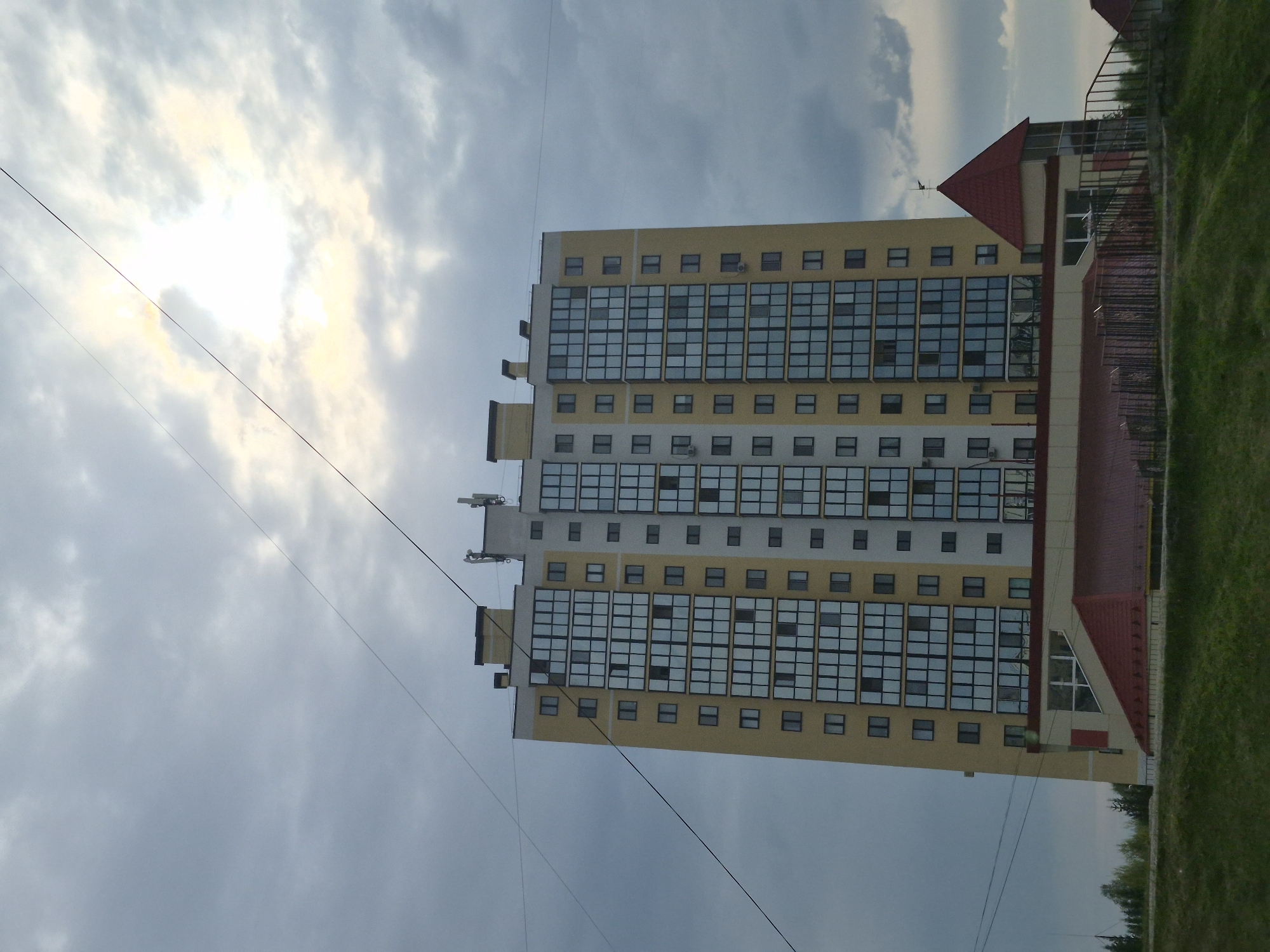 The image shows a high-rise building in Tobolsk, Russia. The building has a yellow facade with rows of rectangular windows. Next to the high-rise is a lower building with a red roof. The sky is overcast with clouds. The ground is covered in grass. Overhead power lines are visible in the sky.