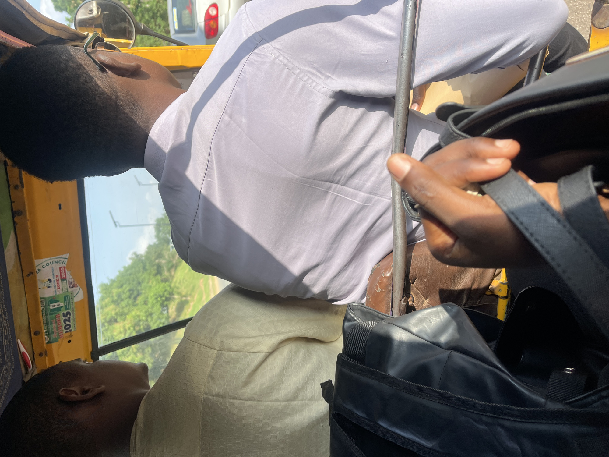 The image shows two individuals inside a vehicle, likely a rickshaw or a similar open-air transport, as indicated by the visible yellow exterior and a mirror. The scene appears to be outdoors during the daytime, with bright sunlight casting shadows and highlighting textures. The overall atmosphere suggests a journey or commute.
In the foreground, a person's hand is visible, holding onto a black bag. The hand has dark skin tone. The black bag is open, revealing a dark interior. Behind this, a person wearing a light-colored, possibly white or light grey, long-sleeved shirt is seated. The shirt is slightly wrinkled, and sunlight illuminates its fabric. The person's back and side are visible, with the head turned away from the camera.
In the background, another person is seated, with their head leaning back and eyes closed, suggesting rest or contemplation. This person has dark skin and short hair. They are wearing a light-colored garment, possibly the same as the person in the foreground or a cream-colored patterned fabric. The interior of the vehicle is visible, including the yellow framing and parts of the seats.
Through the window or opening of the vehicle, lush green trees and foliage are visible, indicating a possibly rural or semi-urban setting. The sky is bright, suggesting good weather.
On the side of the vehicle, some stickers are affixed. One sticker appears to be an election-related campaign poster, with text such as "COUNCIL B," "ZONE B," and "2025." Another sticker has text that is partially obscured but includes what might be "NIGERIA 2025." The location context of Galadimawa Village, Nigeria, aligns with the presence of this type of transport and the visual cues of the environment. No specific emotions are clearly discernible from the individuals, but the person with eyes closed might be experiencing a sense of relaxation.