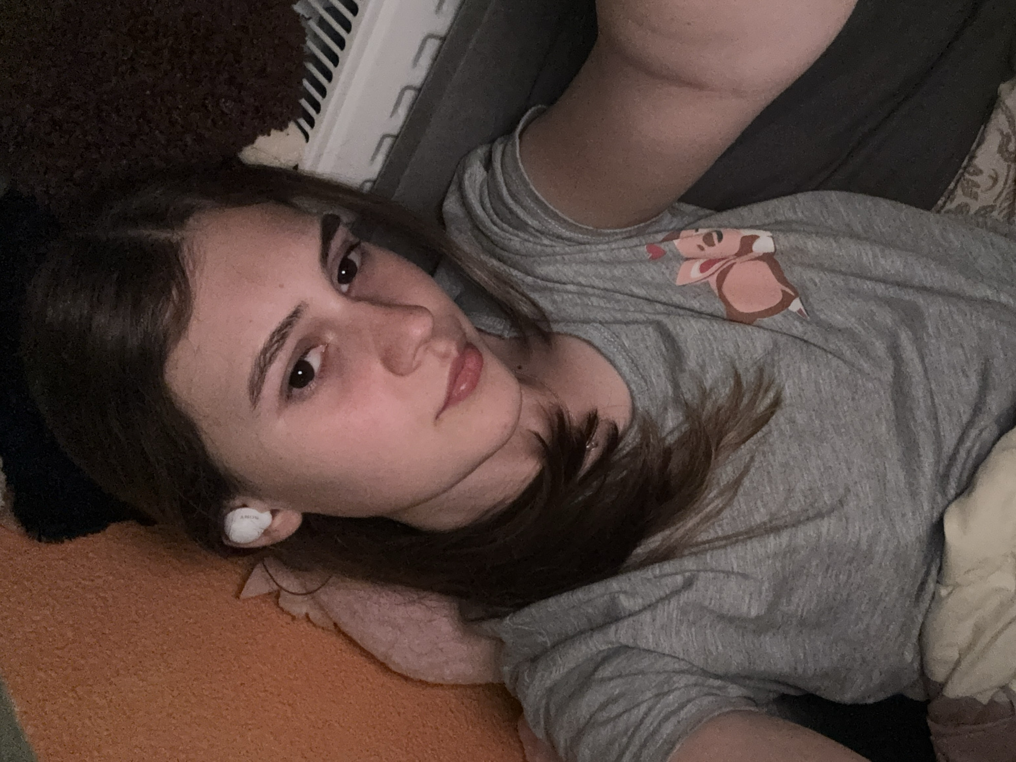 A young person, likely female, is pictured indoors, lying on their left side and looking directly at the camera with a calm, relaxed expression. They have long, dark hair parted on the side and dark eyes. A white SONY earbud is visible in their left ear.
The person is wearing a short-sleeved grey t-shirt, which features a cartoon character resembling a light brown squirrel or chipmunk holding a red heart. They are partially covered by an orange textured blanket or cloth, and dark fabrics, possibly a pillow or blanket, are seen around them. In the upper background, a white object with parallel slats, suggestive of a radiator or window blind, is visible. The lighting is soft, indicating an indoor setting, possibly a bedroom or living room, in Panagyurishte, Bulgaria. No specific activity beyond posing for the photograph and listening to music/audio via the earbud is discernible.