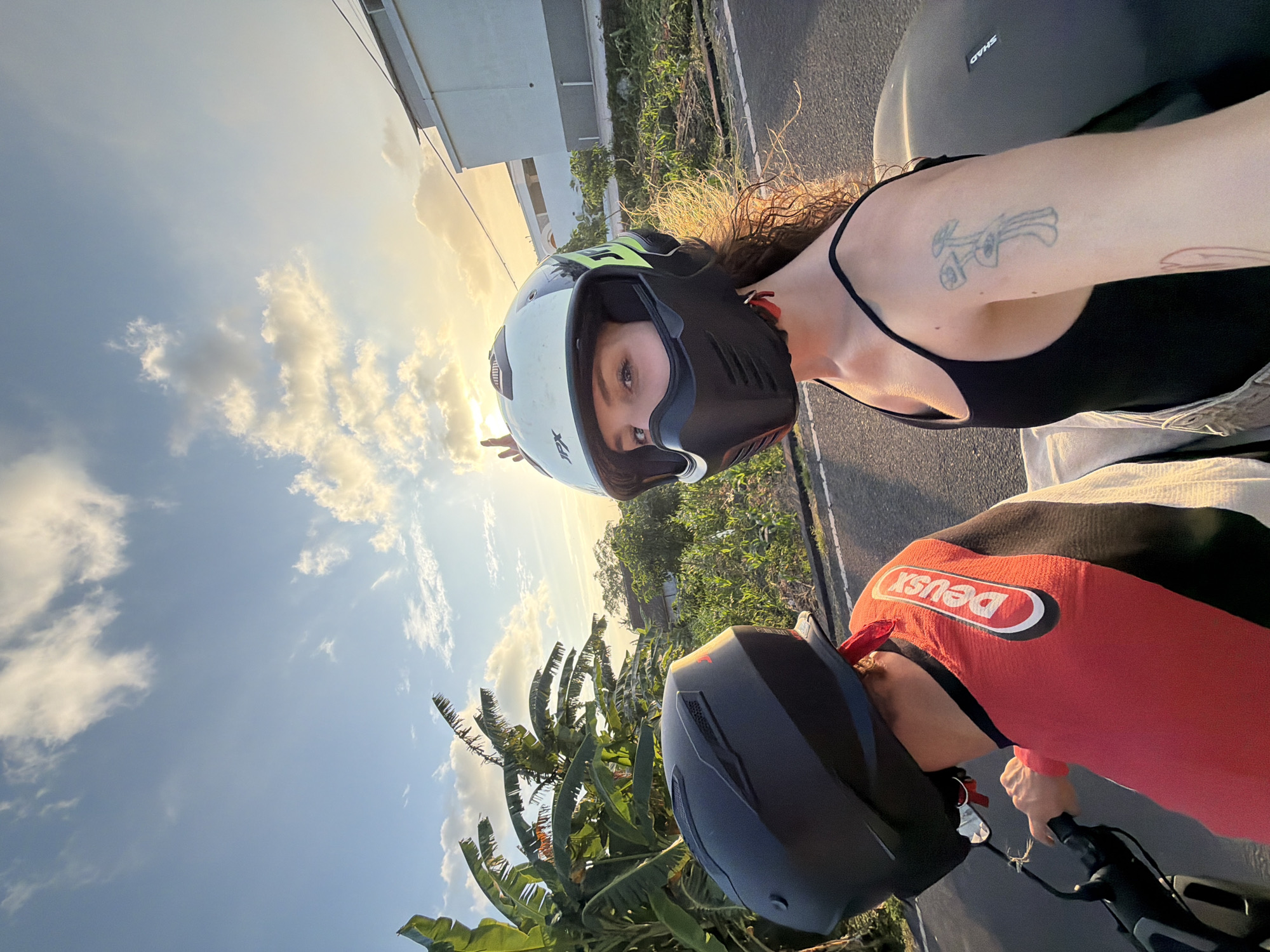 This is a selfie taken from the perspective of a person riding on the back of a motorcycle in North Kuta, Indonesia. The rider in the front is wearing a black helmet and a red and black jacket with the logo "Deus". The person taking the selfie is wearing a white and black helmet with the "JPX" logo, a black tank top, and has curly brown hair. They are looking at the camera and making a peace sign with their left hand. Their right arm is visible, showing a tattoo of three mushrooms and a red heart tattoo. The background shows a road, lush greenery with banana trees, a building, and a cloudy sky with the sun setting. The sun's rays are visible, casting a warm glow on the scene.