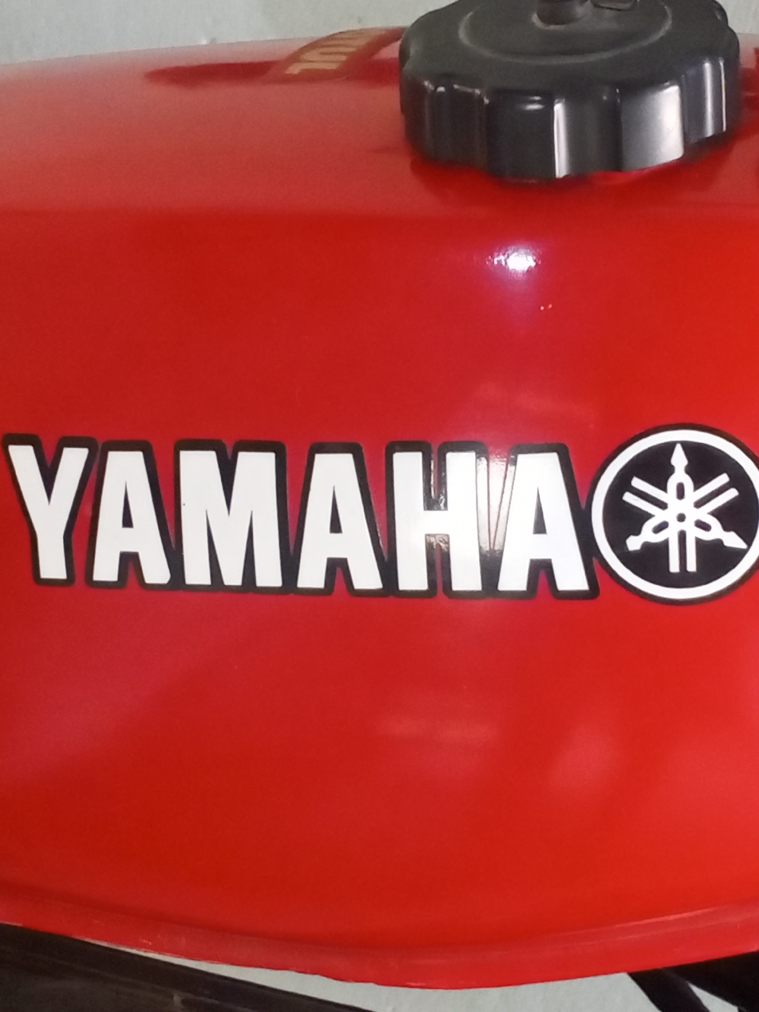 A close-up, eye-level shot shows the side of a red fuel tank from a Yamaha motorcycle. The tank has a glossy finish and is illuminated by overhead light, creating reflections. The word "YAMAHA" is emblazoned in large white block letters with a black outline across the left side of the tank. To the right of the word is the Yamaha logo, a circular emblem with a stylized asterisk design in white on a black background, outlined in white. A black fuel cap with a ridged edge is visible at the top of the tank. The lower edge of the tank curves downwards, revealing a sliver of a dark object, possibly part of the motorcycle's frame or stand, against a white background. The scene is likely an outdoor setting in Altagracia, Venezuela, given the context, though the immediate environment does not offer specific urban cues.