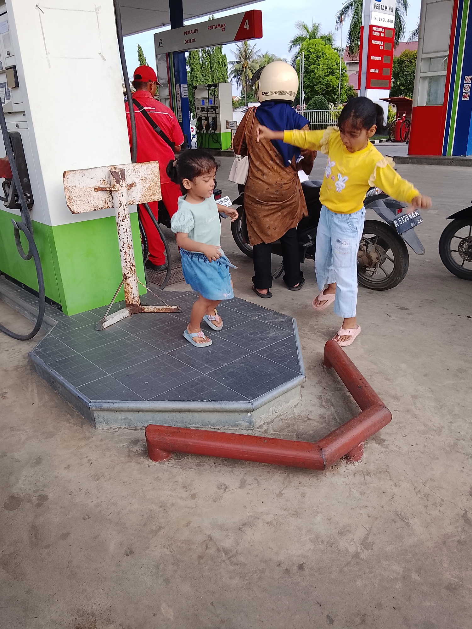 In Lhokseumawe, Indonesia, two young girls are playing at a Pertamina gas station. The girl on the left, wearing a light blue top and a blue skirt, stands on a raised tiled platform in front of a green and white gas pump. The girl on the right, in a yellow long-sleeved shirt and light blue pants, is near a motorcycle, with her arms outstretched as if dancing or playing. A woman in a brown hijab and brown outer garment stands behind her, also near the motorcycle. A gas station attendant in a red uniform is visible behind the younger girl. The scene appears to be taking place in the late afternoon, with overcast skies. Visible text includes "PERTALITE", "DEXLITE", "PERTAMAX 92", "PERTAMINA", and a license plate number "BL 5328 NAK". The overall mood seems playful, with the children engrossed in their activities despite the functional setting of a gas station.