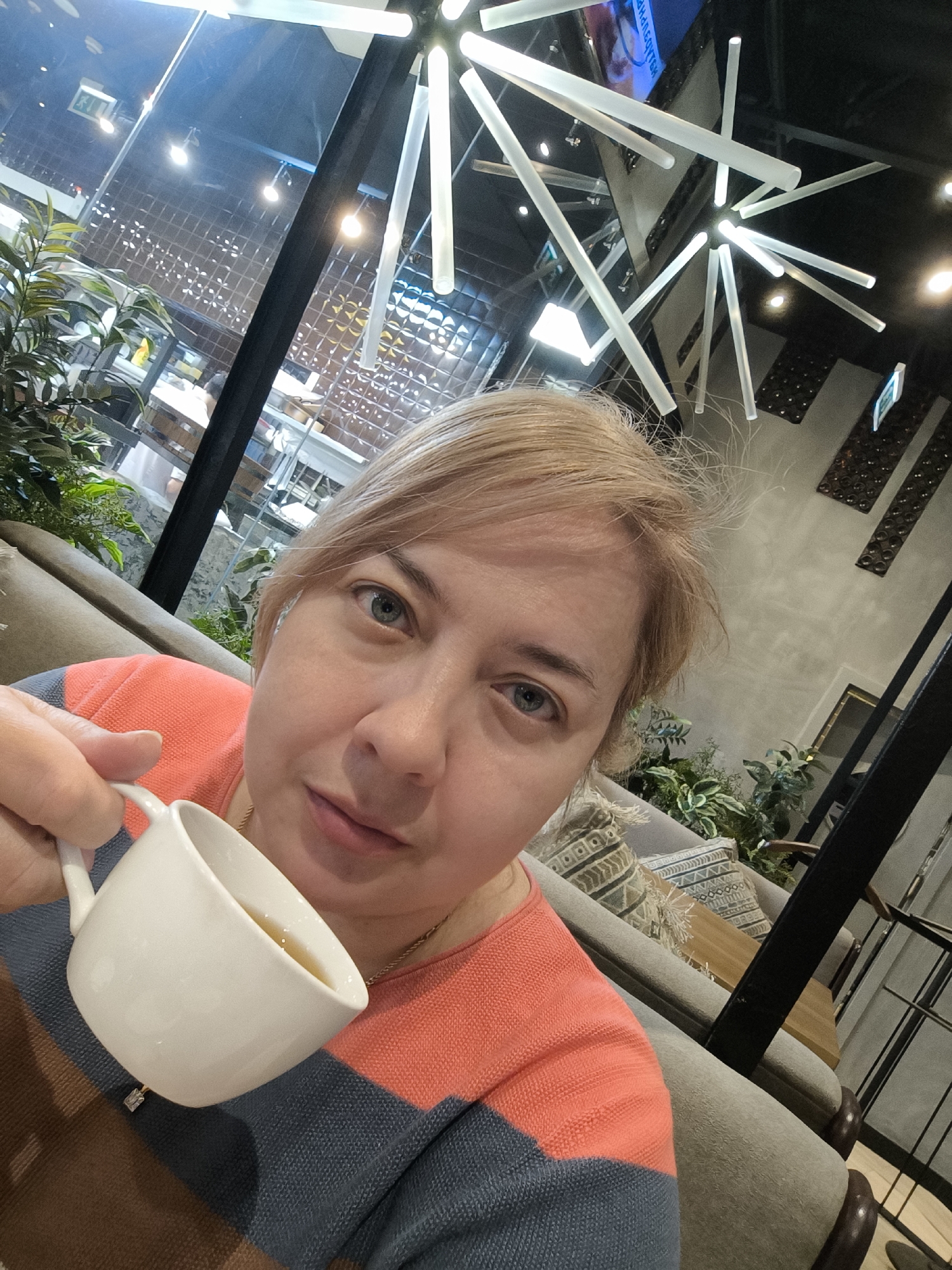 A middle-aged woman with blonde hair holds a white coffee cup in a café. The woman's sweater is striped with coral, blue, and brown. In the background, there are green plants and other seating. Overhead is a modern light fixture consisting of several white, thin cylindrical lights. The reflection in the window behind the woman shows the interior of what appears to be a shopping mall. This image was taken in Khimki, Russia.
