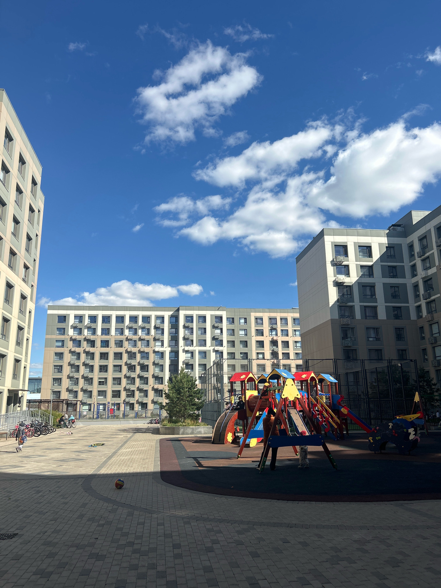 In Astana, Kazakhstan, a residential courtyard is depicted under a bright blue sky with scattered white clouds. The scene features several modern, multi-story apartment buildings with light-colored facades and numerous windows. In the foreground, a children's playground is set up with various colorful structures, including slides, swings, and climbing equipment, situated on a dark rubberized surface. Adjacent to the playground is a paved area made of interlocking gray bricks, where a basketball lies on the ground. Further into the courtyard, a line of parked bicycles is visible, and a few individuals are scattered around, some appearing to be children playing, while others are adults. A small evergreen tree stands near one of the buildings. The overall impression is of a well-maintained, contemporary urban living space designed for families.