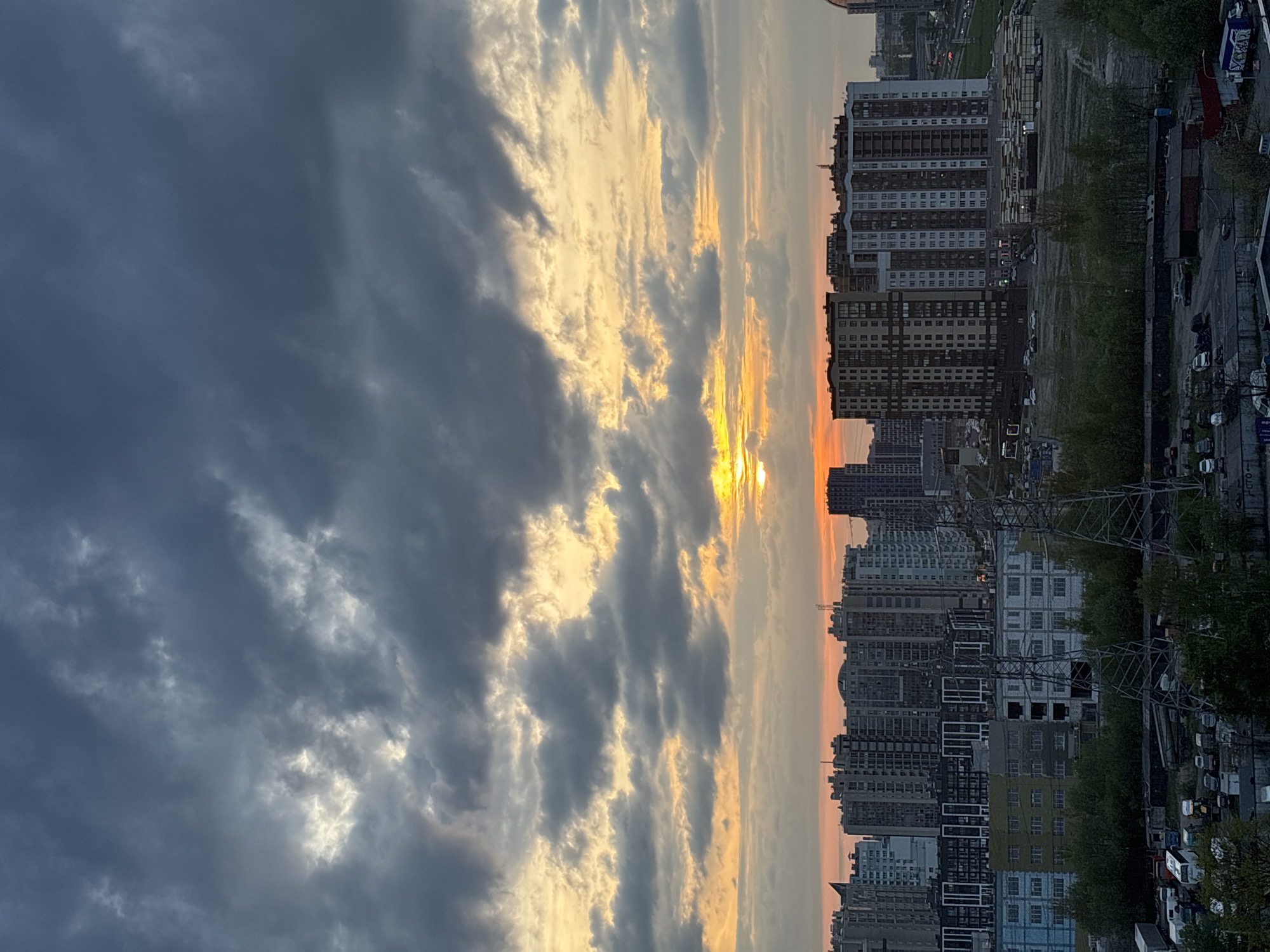 The image captures a cityscape at sunset. Buildings of varying heights and designs line the horizon, with a mix of residential and commercial structures. A road runs alongside the buildings, populated with cars. Overhead power lines and other infrastructure are visible. The sky is filled with clouds, with the sun setting between them, casting a golden light. The general location appears to be a dense urban area, possibly a large city.