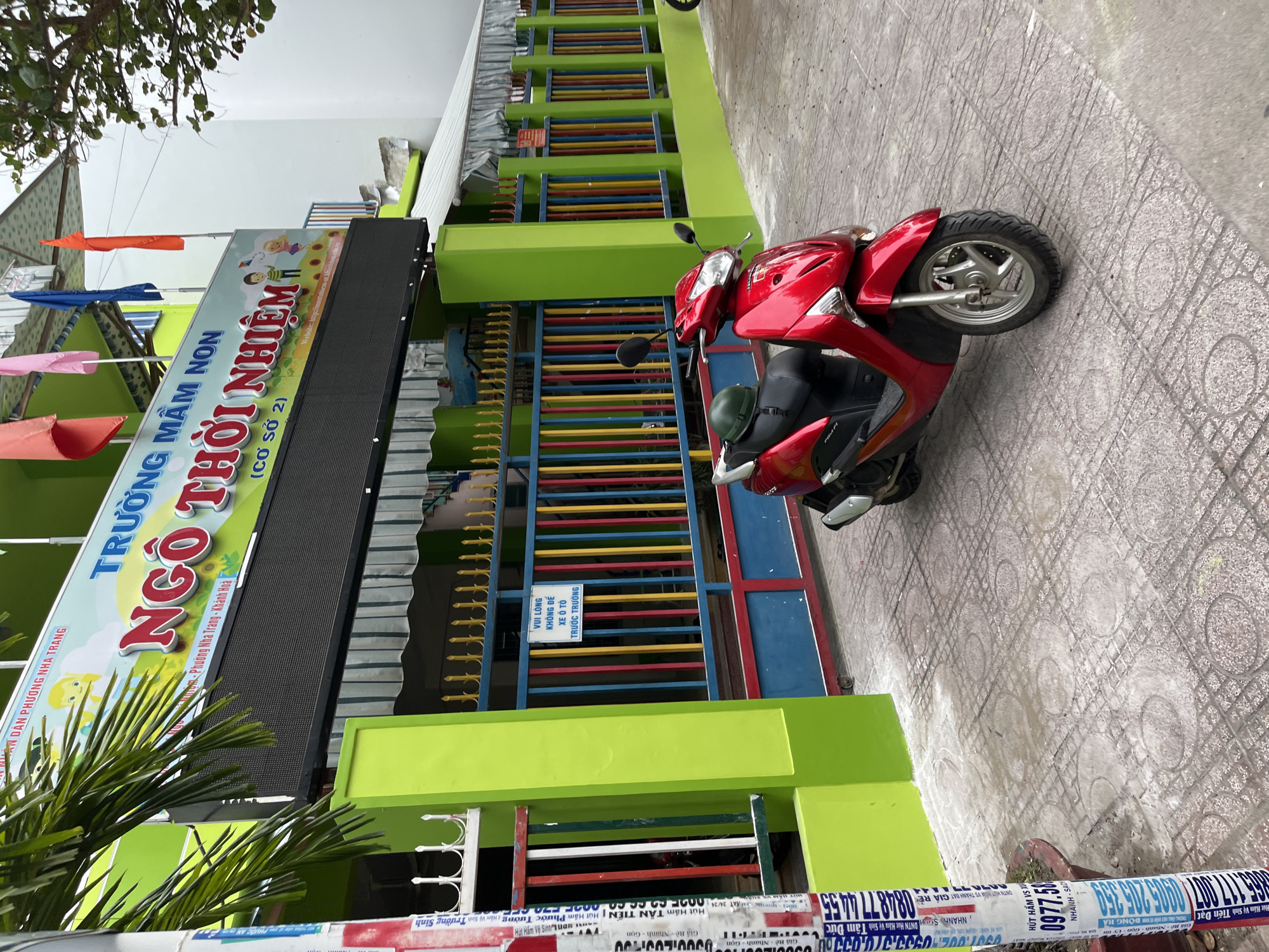 The image shows the entrance to a kindergarten in Nha Trang, Vietnam. A bright green building with a colorful fence in front of it is visible. A red scooter is parked on the sidewalk in front of the fence. The kindergarten's name, "TRƯỜNG MẦM NON NGÔ THỜI NHIỆM (CƠ SỞ 2)", is displayed on a banner above the entrance. The fence is made of vertical red, yellow, blue, and green bars. A sign on the fence reads "VUI LÒNG KHÔNG ĐỂ XE Ô TÔ TRƯỚC TRƯỜNG", which translates to "Please do not park cars in front of the school." There is a palm tree on the left side of the image, and a white building in the background. The ground is paved with tiles. There are some advertisements on a pole to the left of the entrance.