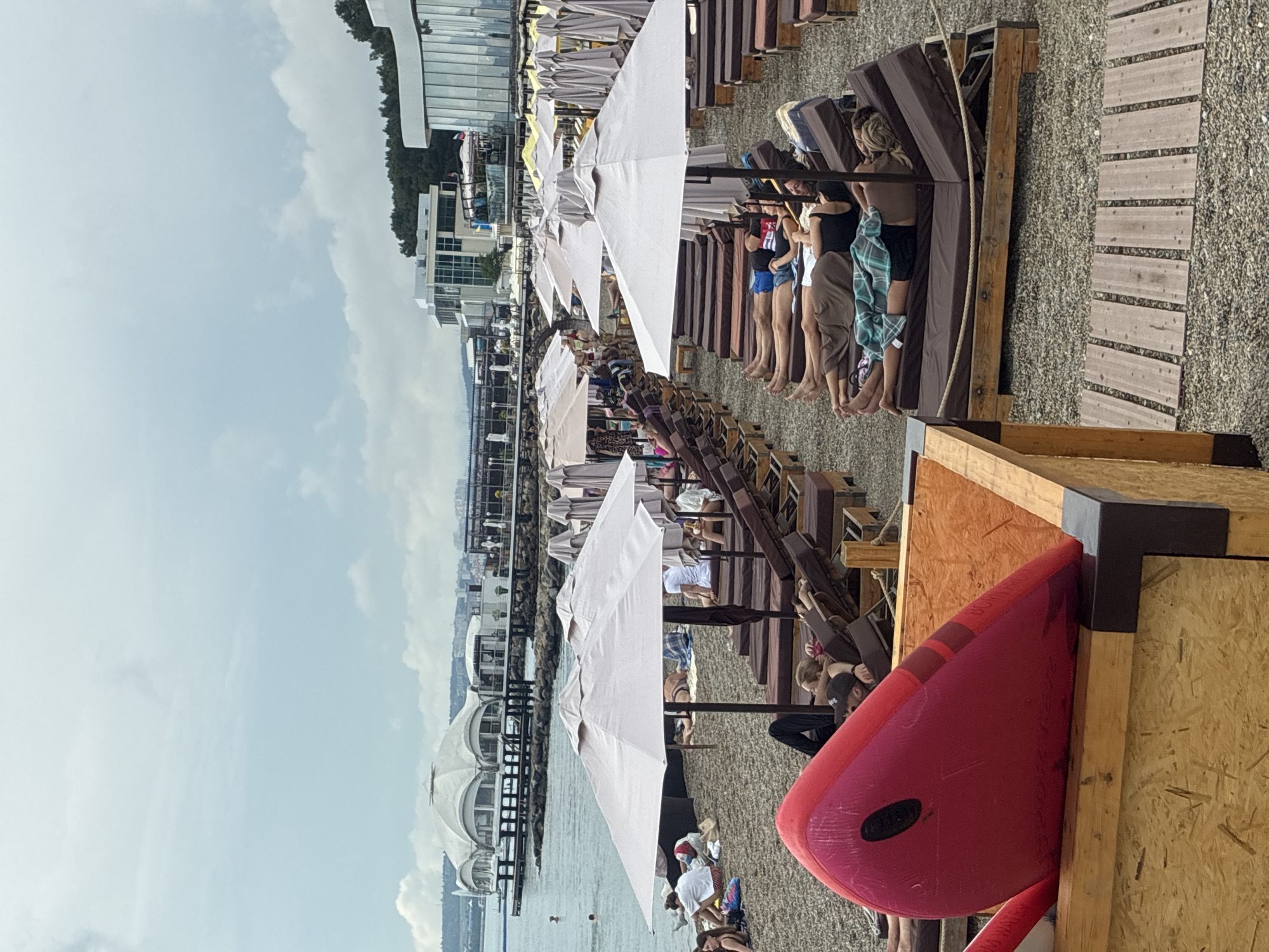 The image depicts a crowded beach in Gelendzhik, Russia, characterized by rows of unoccupied and occupied sun loungers covered by umbrellas. In the foreground, a bright pink inflatable object is placed inside a wooden container. The beach is composed of pebbles, and a wooden walkway is visible in the lower right portion of the frame. In the background, several modern buildings are situated along the coastline, with a distinctive white domed structure on a pier extending into the water. The sky is overcast. People are seen relaxing on the sun loungers and a few individuals are in the water. The overall scene conveys a typical summer day at a resort beach.