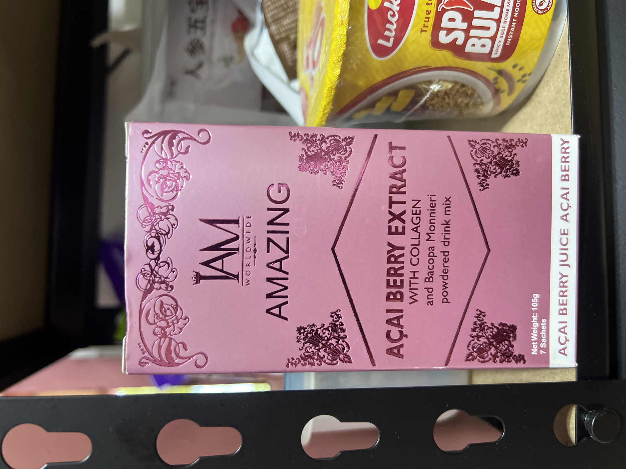 The image shows a box of "I AM AMAZING" Acai Berry Extract powdered drink mix with collagen and Bacopa Monnieri. The box is pink with decorative floral patterns and text in a serif font. The product is labeled as being "Worldwide." The box has a net weight of 105g and contains 7 sachets. Behind the box, a package of "Lucky Me! Spicy Bulalo" instant noodles is visible. The box is placed on a black metal shelf, suggesting it is inside a cabinet or storage unit. The location in Davao City, Philippines, cannot be verified from the image.