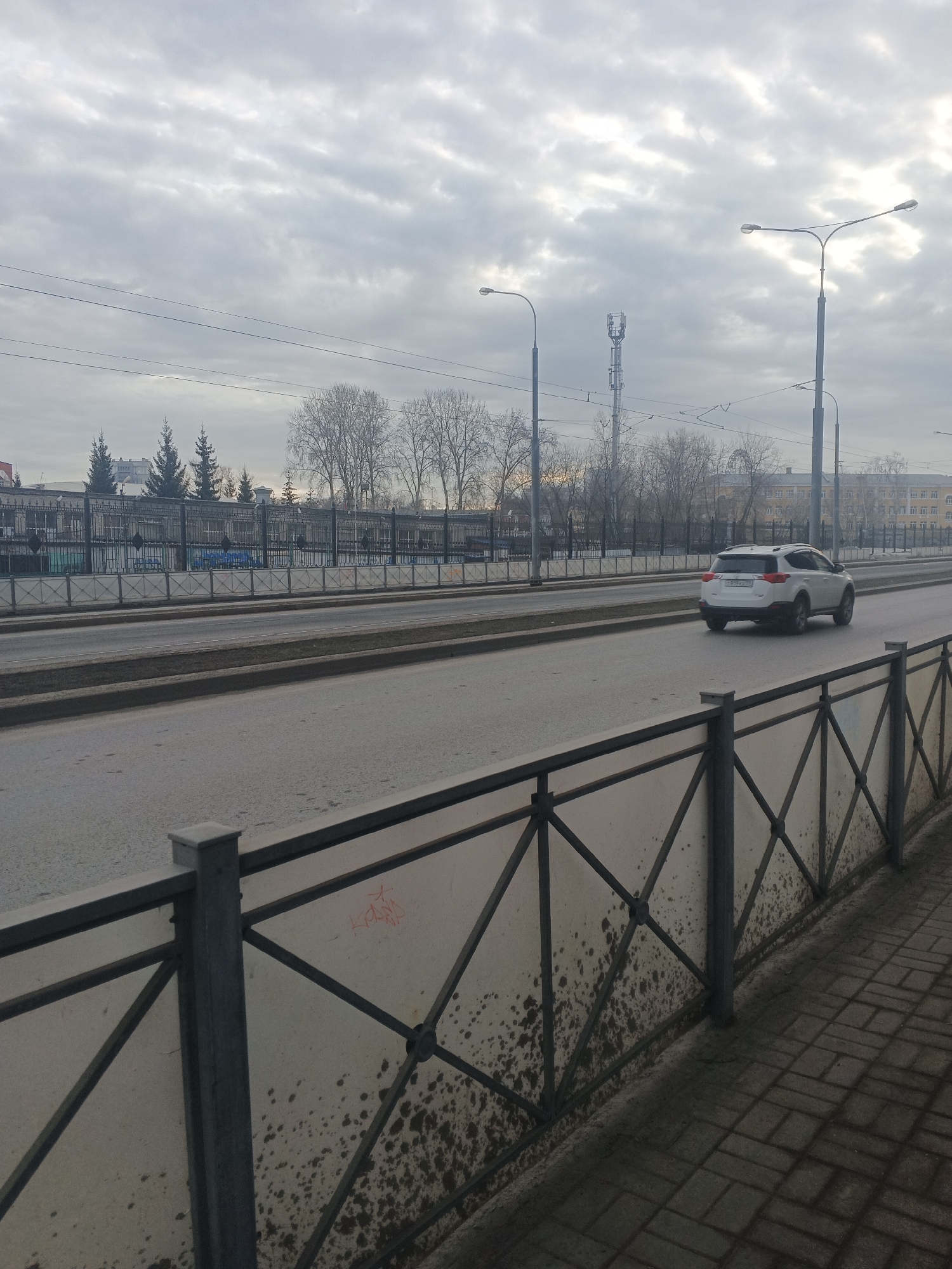 The photograph depicts a street scene in Perm, Russia, on an overcast day. A white SUV is driving on a wide, multi-lane road. The road is bordered by metal fencing on either side. The foreground fencing is marked with dark stains and graffiti. Behind the road, a taller metal fence runs in front of a building complex. Streetlights are visible above the trees. A cell tower rises above the buildings. The sky is filled with grey clouds. The setting suggests an urban environment.