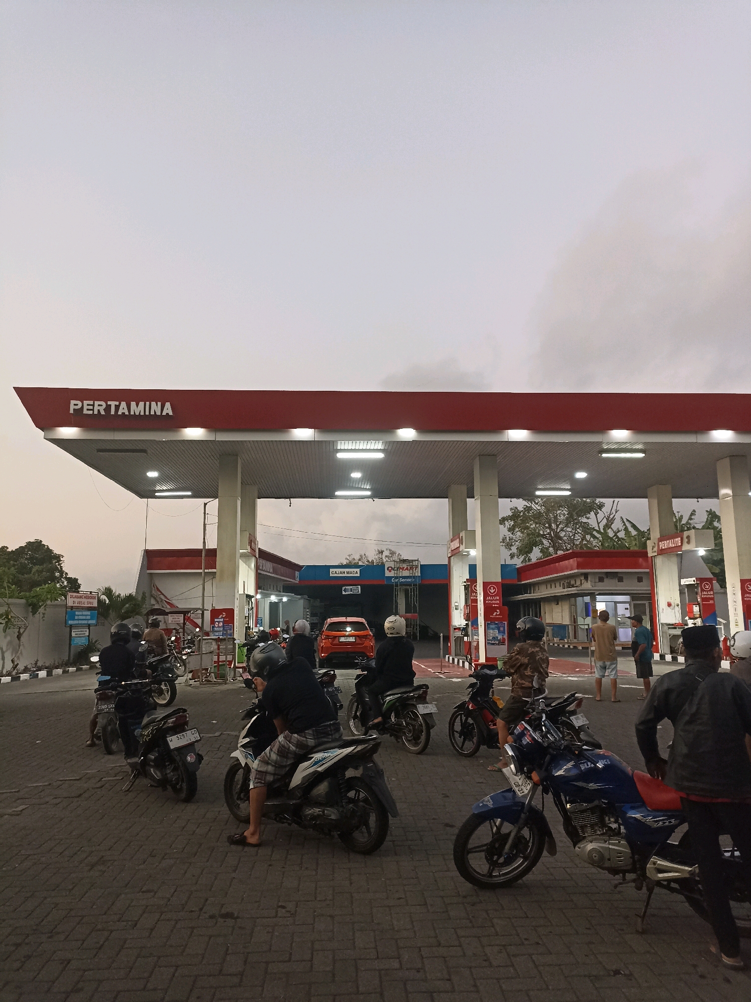 A busy Pertamina gas station in Banyuwangi, Indonesia, is captured in this twilight scene. The prominent red and white Pertamina branding is visible on the canopy overhead. Several motorcycles, some with riders waiting, are parked on the brick-paved lot. One orange car is visible at a pump. People are standing and walking around the station. The sky is overcast, suggesting a cloudy day or evening. The overall atmosphere is one of typical activity at a public service station.