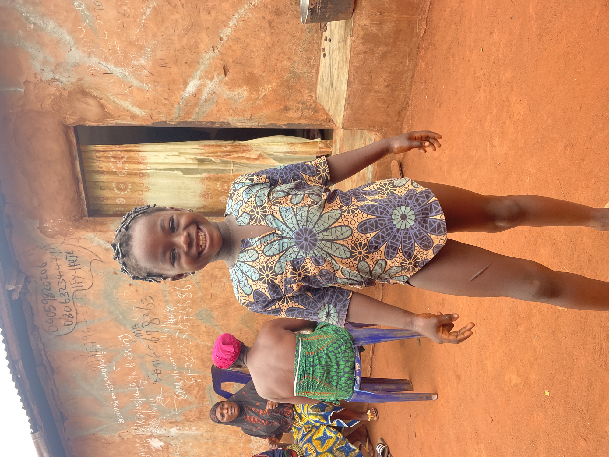 A medium shot shows a girl standing in front of a building with writing on the wall. She has dark skin, short dark braids, and is wearing a printed dress with blue, yellow, and black floral patterns. She is smiling, and her arms are outstretched. Behind her and to the left, there is an adult seated on a blue plastic chair with their back to the camera, wearing a green and white patterned cloth around their waist. To the side of this individual is another seated woman wearing a patterned dress and headscarf. The wall behind them is made of reddish-brown material and has white writing scrawled on it, including phone numbers and names. The ground is orange-red earth. The image appears to have been taken outdoors, possibly in the city of Ogbe, Nigeria. A metal bucket hangs from the roof.