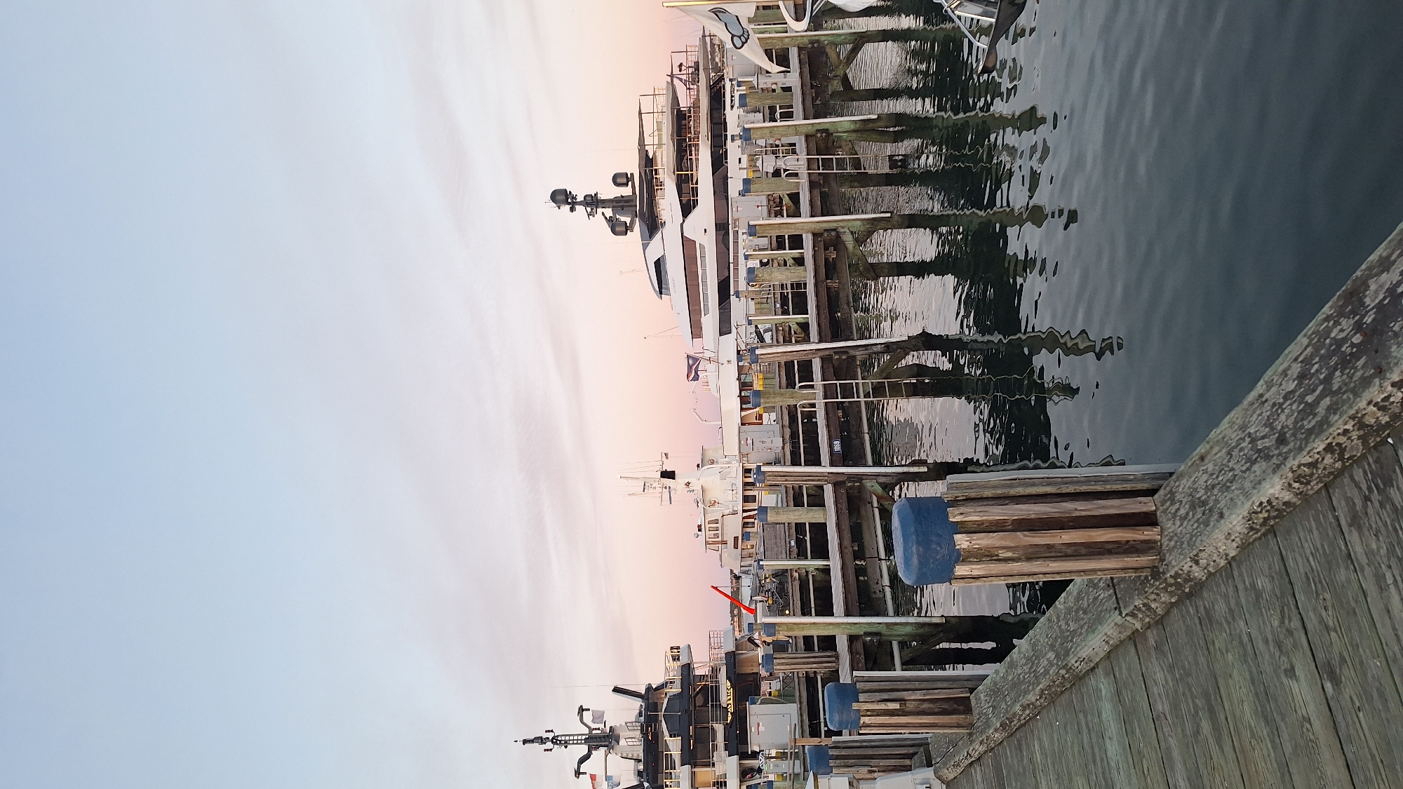 The image depicts a serene harbor scene in Nantucket, United States of America, at dusk. In the foreground, weathered wooden planks form a pier, leading the eye towards the water. Several mooring posts, some topped with blue caps, punctuate the pier's edge. Beyond, a fleet of luxurious yachts is docked.
The most prominent vessel on the left is a black and white yacht with a tall mast. To its right, a smaller white boat with a tower stands. Further to the right, a large, sleek white yacht dominates the scene, its modern design contrasting with the rustic pier. The yachts are moored to a wooden dock structure.
The water in the harbor is calm, reflecting the structures and the soft hues of the twilight sky. The sky itself transitions from a pale blue at the top to a gentle pink and orange near the horizon, indicative of a sunset. The overall atmosphere is one of tranquility and maritime elegance. There are no people visible in the image, emphasizing the quiet stillness of the harbor at this time of day. The lighting suggests late afternoon or early evening.
