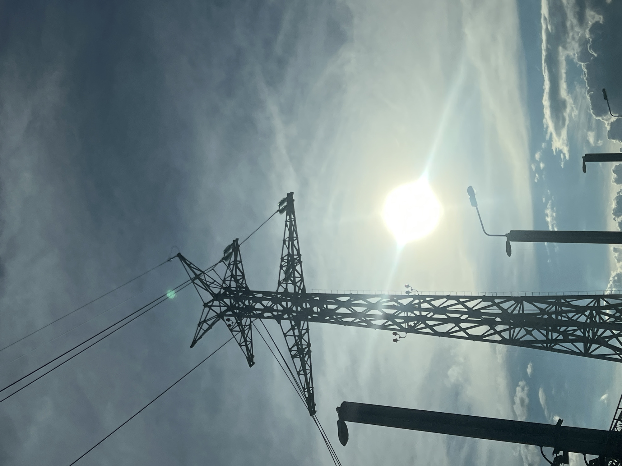 A low-angle shot depicts an electricity pylon in Chaikovsky, Russia, silhouetted against a sky with scattered clouds and a bright sun. The metal framework of the pylon is prominently visible, including the insulators that attach the power lines to the structure. To the right of the pylon, two streetlights are visible.