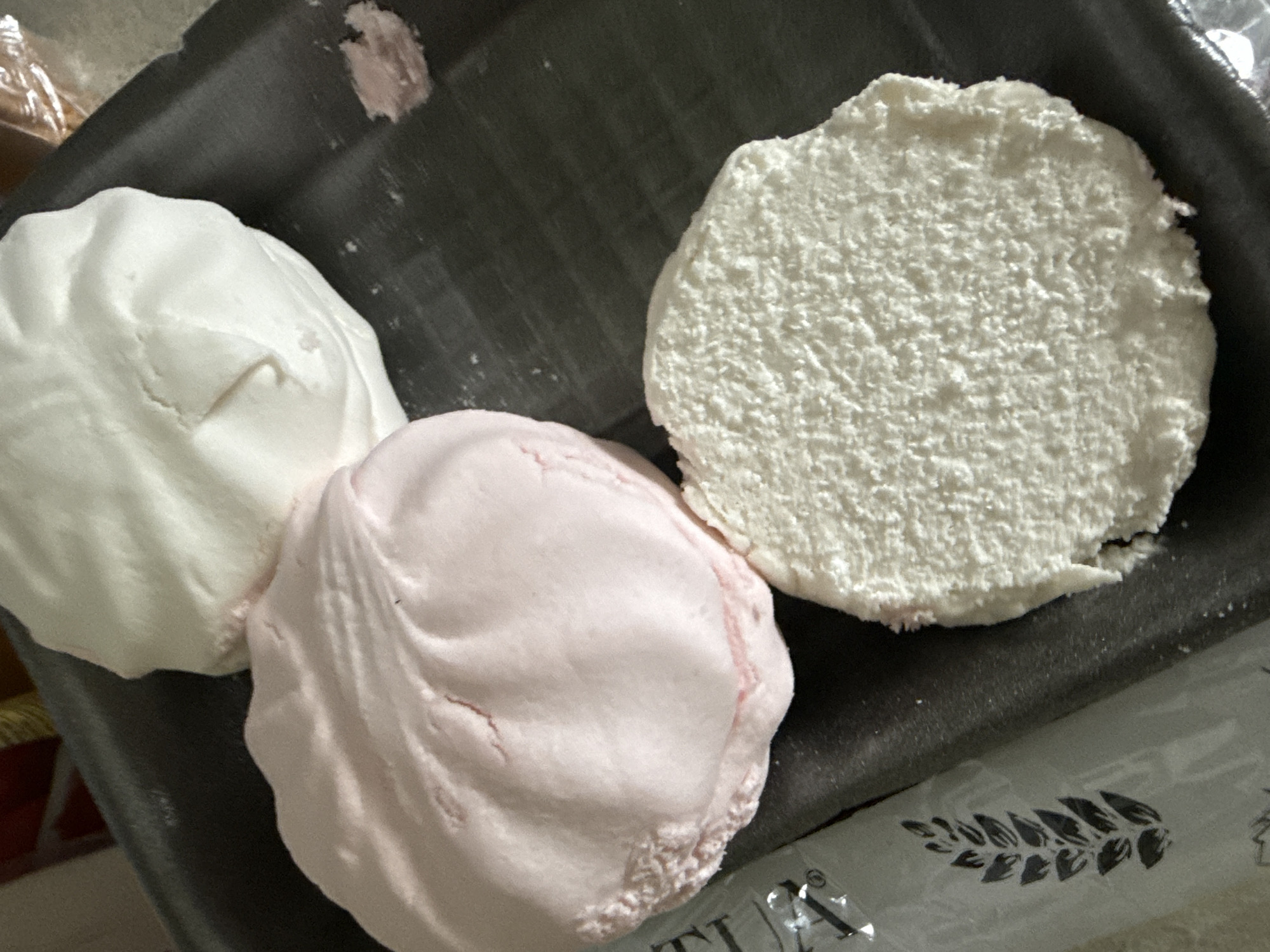 Three mounds of dough, one pink and two white, are placed on a dark grey plastic tray. The dough appears to be uncooked, with the pink mound showing visible indentations and streaks. One white mound is smooth and rounded, while the other is flattened and has a textured, grainy surface, possibly from being dusted with flour or sugar. A small piece of pink dough has broken off and is visible on the tray. To the left of the tray, a portion of a silver wrapper with a black botanical print is visible. The background is blurred, but suggests an indoor setting with red and yellow objects. There are no people or actions depicted. The image does not provide any information about the location in the city of Okhtyrka, Ukraine.