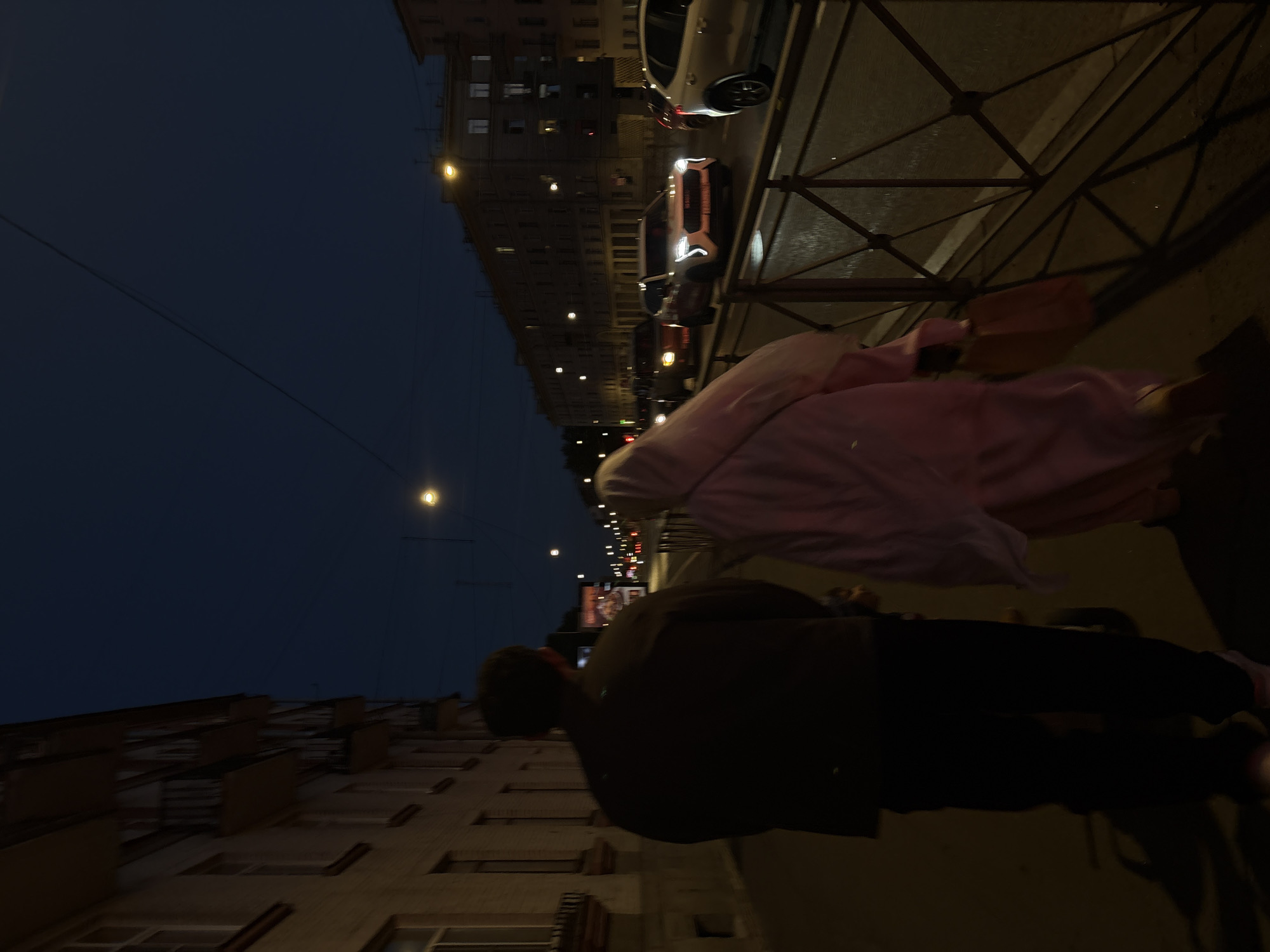 Two individuals, a male from the rear and a female in a pink outfit, walk along a sidewalk in Saint Petersburg, Russia, at dusk. The male is dressed in a dark jacket and dark pants. The female is wearing a light pink hooded garment and carrying a bag in her right hand. They are walking on a sidewalk adjacent to a street with traffic, including several cars. The street is lined with apartment buildings on the right side of the image, illuminated by streetlights and lights from the building windows. A metal railing separates the sidewalk from the street. The sky is dark blue, indicating evening. The building on the left is a multi-story structure with balconies. The overall scene depicts a typical urban environment in Saint Petersburg during the evening hours.