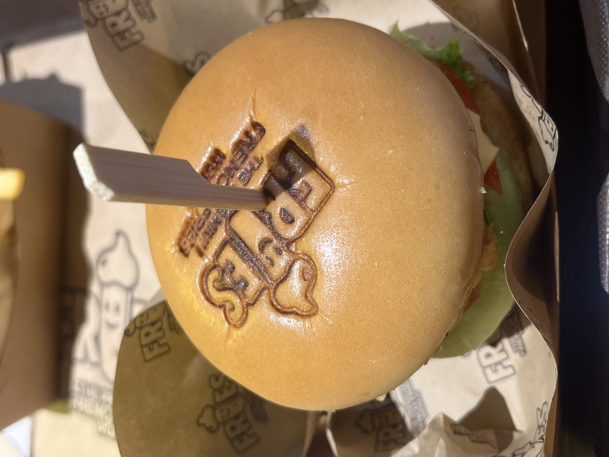 A top-down shot of a burger in Thủ Đức, Vietnam. The burger bun has the "FRY'S" logo branded on top and is held together by a wooden pick. The burger is placed in a paper wrapping, also branded with "FRY'S." Part of the burger's contents, including lettuce and tomato, are visible. The burger is situated in its serving container.