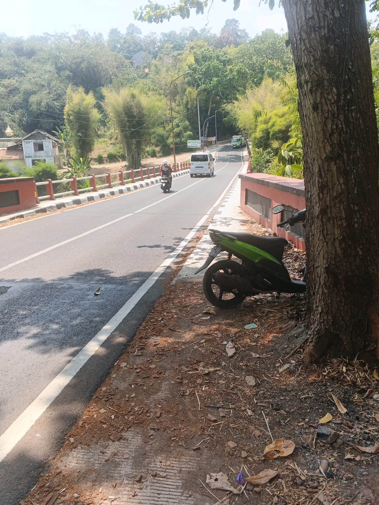 A scenic, sun-drenched road winds through a lush, green hillside in Semarang, Indonesia. A single-lane road, marked with a white line, curves to the left, bordered by a concrete barrier with red and white stripes. On the right side of the road, a green motorcycle is parked next to a large tree. A few leaves and debris are scattered on the ground.
In the distance, a white van drives away from the camera, and a green truck is visible further up the road. A person on a motorcycle heads towards the camera. The hillside is covered in dense vegetation, including tall bamboo stalks and various trees. A small, light-colored building with a red roof is visible on the left side of the road, near a white sign that reads "WATERFALL." The sky is bright and clear, suggesting a sunny day. The overall scene depicts a rural or semi-rural road environment in Semarang, Indonesia, characterized by natural beauty and the presence of vehicles.