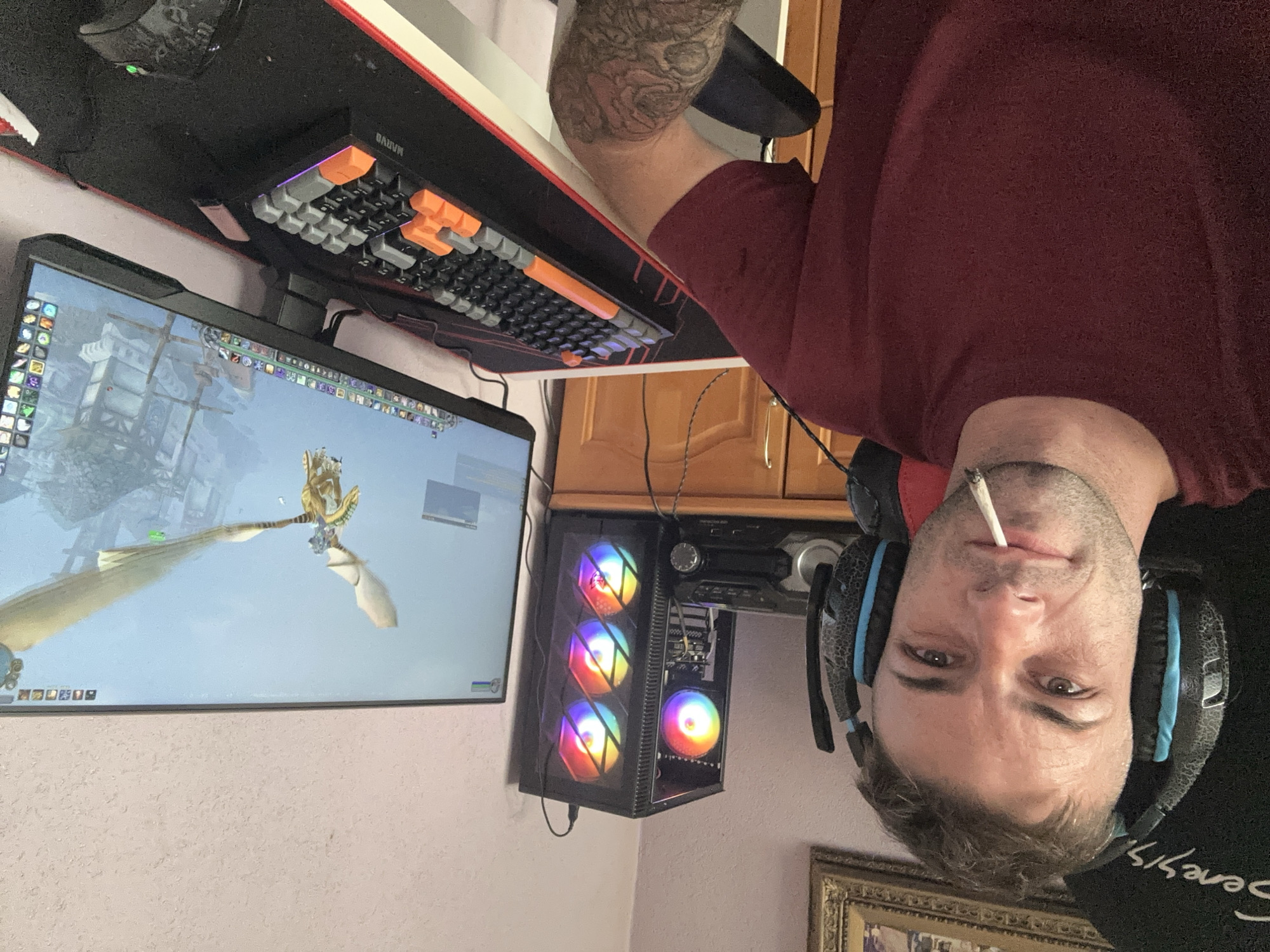 The image shows a Caucasian male in his late 20s or early 30s, with short brown hair and a light beard, wearing a maroon t-shirt and large headphones. He is seated in a gaming chair, with a cigarette in his mouth. To his right, there is a computer tower with RGB lighting, a stereo system, and a large monitor displaying a video game. A gaming keyboard and mouse are on a desk in front of him. The environment appears to be a home office or gaming room, with wooden furniture and a framed picture on the wall. The image does not provide any specific information to determine the location within the city of Cherven Breg, Bulgaria.