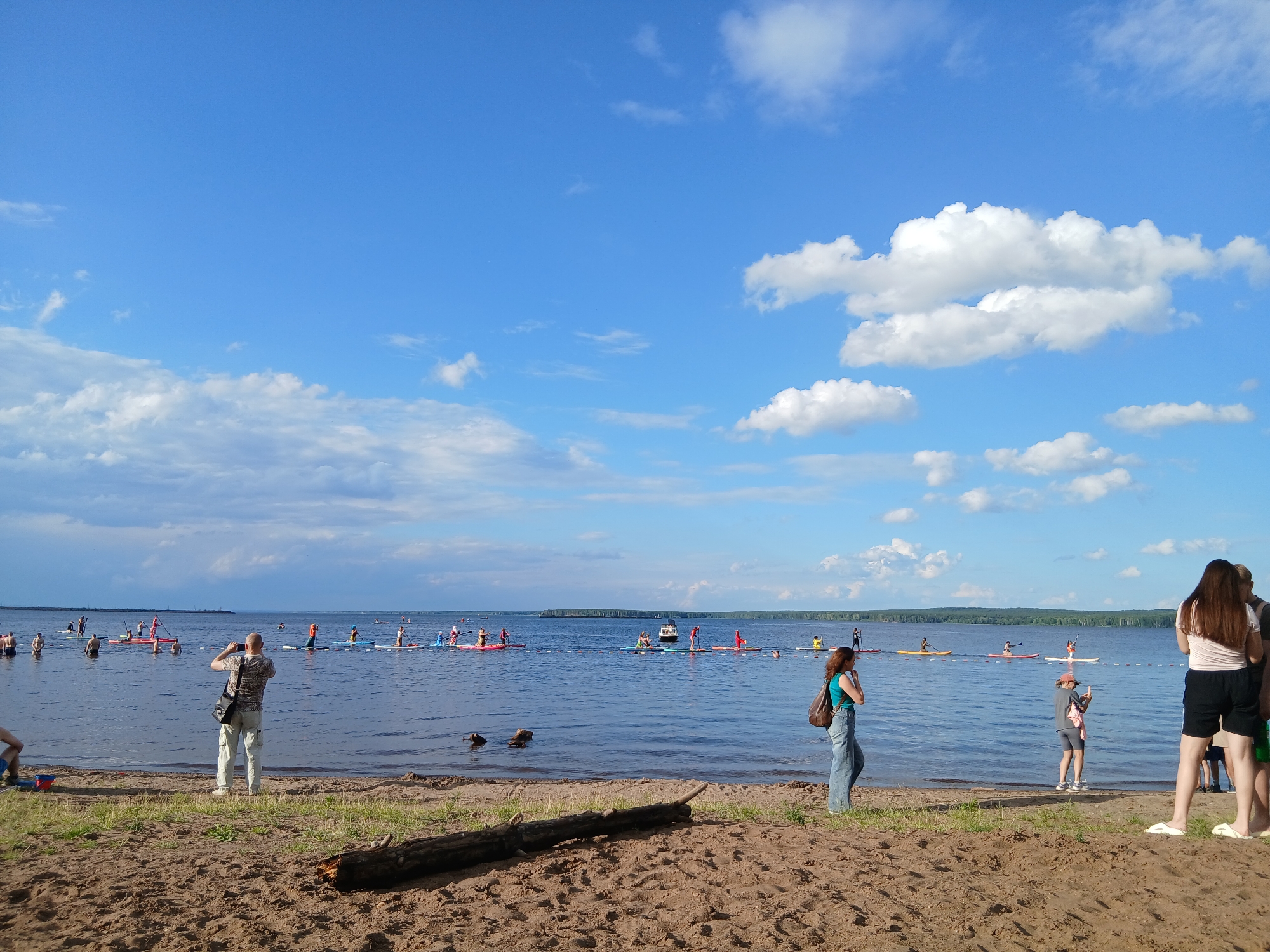 The image depicts a sunny day at a beach in Chaikovsky, Russia. The scene is filled with people enjoying various activities on and near the water. In the foreground, a sandy beach is visible with a weathered log lying on it. A few individuals are present on the beach, including a person taking a photo with a phone and a woman with a backpack.
The main focus of the image is the water, which is dotted with numerous people on paddleboards. They are spread out across the calm surface of a large body of water, possibly a lake or river. Some are paddling, while others appear to be engaged in conversation or resting. The activity suggests a recreational event or a group outing.
In the distance, a treeline marks the edge of the land, and the sky is a vibrant blue with scattered white clouds, indicating pleasant weather. The overall atmosphere appears relaxed and cheerful, with people taking advantage of the outdoor setting. The time of day seems to be late afternoon, judging by the quality of light. No visible text is present in the image.