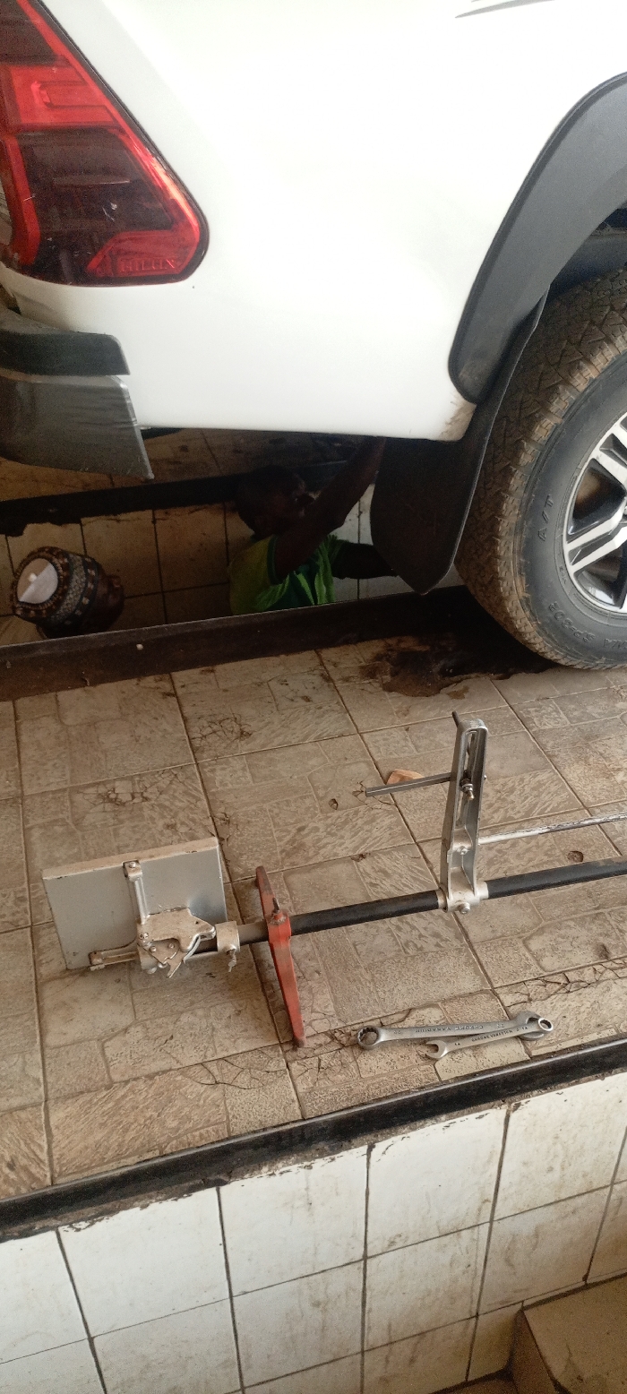 In a vehicle service bay in Kurudu, Nigeria, the rear section of a white Toyota Hilux pickup truck is positioned over an inspection pit. A dark-skinned individual, wearing a green shirt, is visible inside the pit, actively working on the underside of the vehicle. To the individual's left, an ornamental, patterned pot rests on the pit floor. The service bay floor is tiled with light brown/beige square tiles, showing signs of dirt and minor cracks. On this tiled surface, near the vehicle's right rear wheel, a silver-colored wheel alignment tool consisting of a central black bar, a rectangular silver measuring plate with a clamp, and a red support, is laid out. Adjacent to the alignment tool, a silver-colored combination wrench (marked "CHROME VANADIUM 22 24") is also on the floor. Dark liquid stains are present on the tiled floor directly beneath the vehicle's right rear axle area. The pit walls are lined with white square tiles.