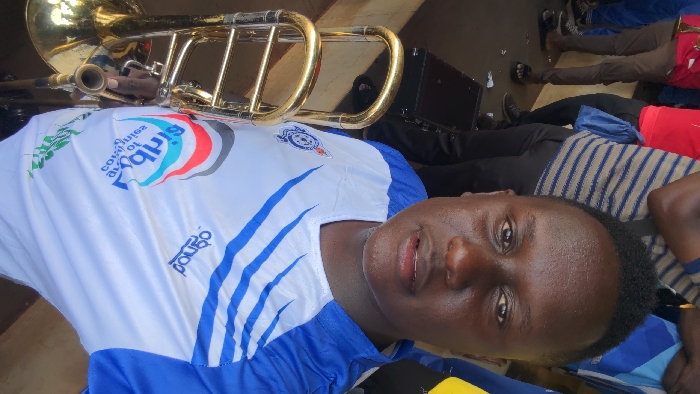 A man is seen wearing a blue and white jersey with sponsor logos. He is partially visible, as he is laying down, angled towards the camera. A gold trombone is positioned above him, possibly being held by another person whose hand is barely visible. Other individuals, some with bags, are seen lying down nearby. The scene appears relaxed and informal.