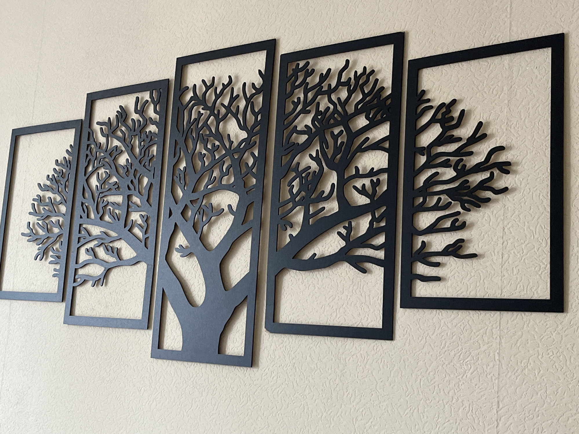 This is a photograph of a wall art installation depicting stylized trees. The installation consists of five black, rectangular panels of varying widths, arranged horizontally. Each panel features an intricate, cutout silhouette of a tree with branching limbs. The background is a textured, light-colored wall. The artwork is displayed indoors, likely in a residential or commercial setting, though its specific location in Odintsovo, Russia, cannot be determined from the image alone. There are no people or animals visible in the scene, and no actions are taking place.