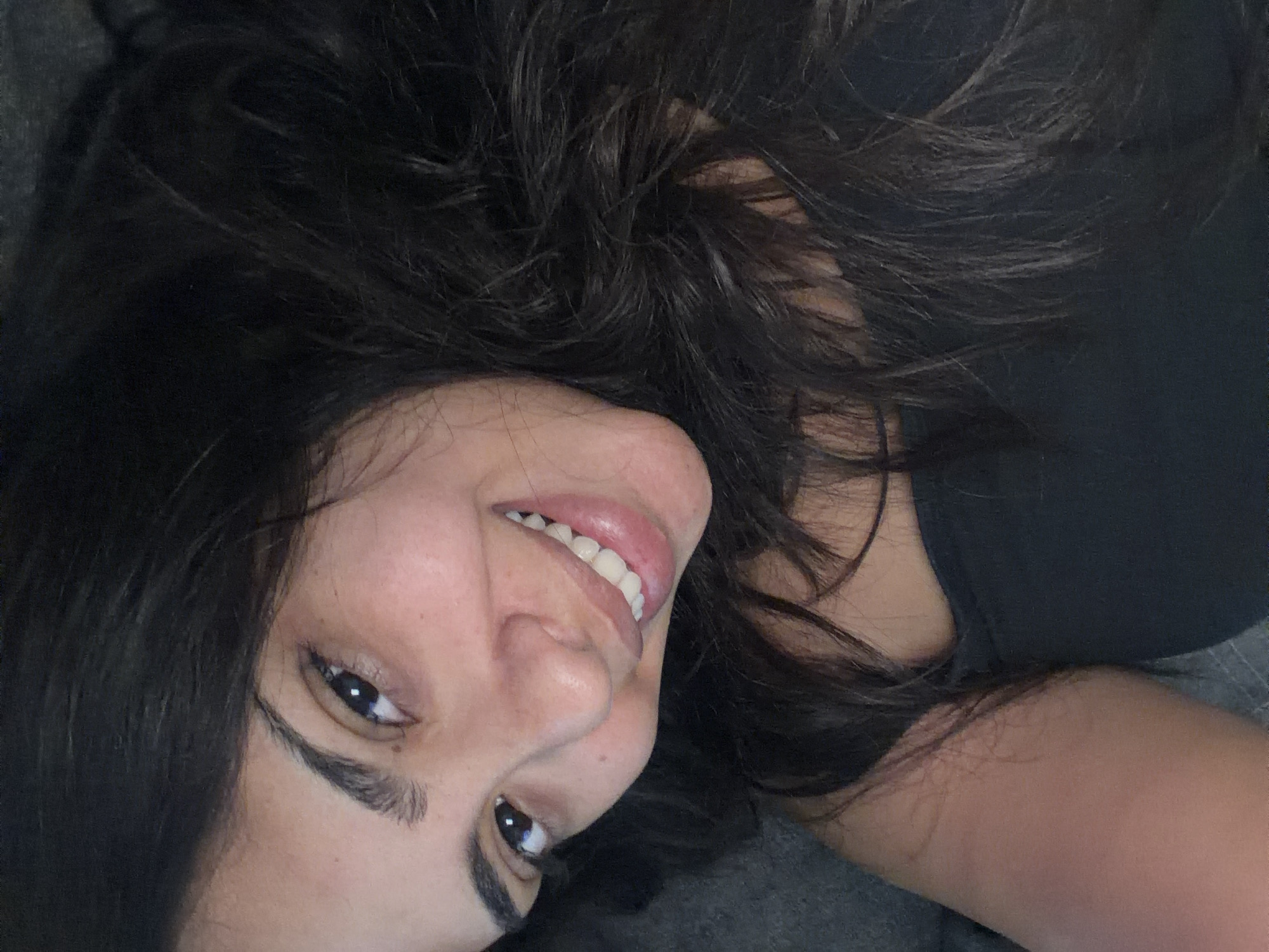 The image is a close-up, selfie-style photograph of a young woman. She has dark, wavy hair that frames her face. Her left eye is clearly visible, looking directly at the camera, with her right eye partially obscured by her hair. She is smiling, revealing white teeth. She is wearing a black strapless top. The background appears to be a dark, textured surface, possibly a piece of furniture or bedding. The lighting is soft and appears to be coming from the front, illuminating her face. There is no discernible context or location within the city of Duisburg, Germany, or any other specific geographical location, evident in the image.
