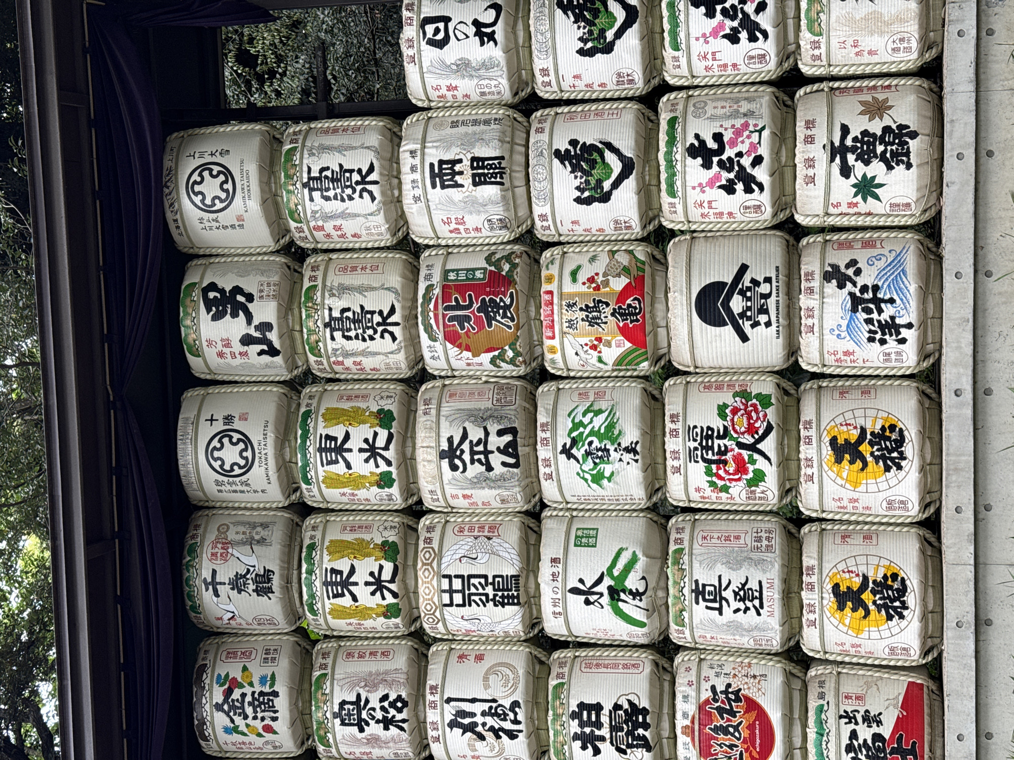This image captures a vibrant display of traditional Japanese sake barrels, known as *kagami-biraki* barrels or *komodaru*, stacked neatly in horizontal rows. There are at least five visible rows and eight columns of barrels, filling the majority of the frame.
Each barrel is cylindrical, wrapped in a straw mat (komo) and secured with rope, featuring a distinct paper label (noren) on its front. These labels showcase a rich variety of calligraphic brand names, traditional Japanese art, logos, and crests, often in black ink with highlights of red, green, blue, yellow, and pink. Many labels also include the term "清酒" (Seishu - refined sake) and "登録商標" (Tōroku Shōhyō - Registered Trademark).
Notable examples of visible text and imagery include "上川大雪" (Kamikawa Taisetsu) from Hokkaido with a circular crest, "男山" (Otokoyama), "高清水" (Takashimizu), "千歳鶴" (Chitose Tsuru), "白鶴" (Hakutsuru), "真澄" (Masumi), "越後桜" (Echigozakura) from Niigata, and "島根" (Shimane), among many others, representing various breweries across Japan. Some barrels display intricate designs like cranes, cherry blossoms, peonies, dragons, or stylized kanji characters. English text such as "KAMIKAWA TAISETSU HOKKAIDO" and "ILAKA JAPANESE SAKE ATELIER" is also present on some labels.
The barrels are displayed outdoors, possibly under a covered structure or an overhang, indicated by the dark, draped fabric and a structural beam on the left, and a concrete divider on the right. Green foliage from trees is visible in the background on the left, suggesting a natural setting. The lighting is soft and even, indicating daytime, possibly overcast conditions or shade. This arrangement is typical of sake offerings displayed at Shinto shrines or temples, often seen at locations like Meiji Jingu in Shibuya, Japan, to honor deities and celebrate the breweries. The display evokes a sense of tradition and cultural heritage.