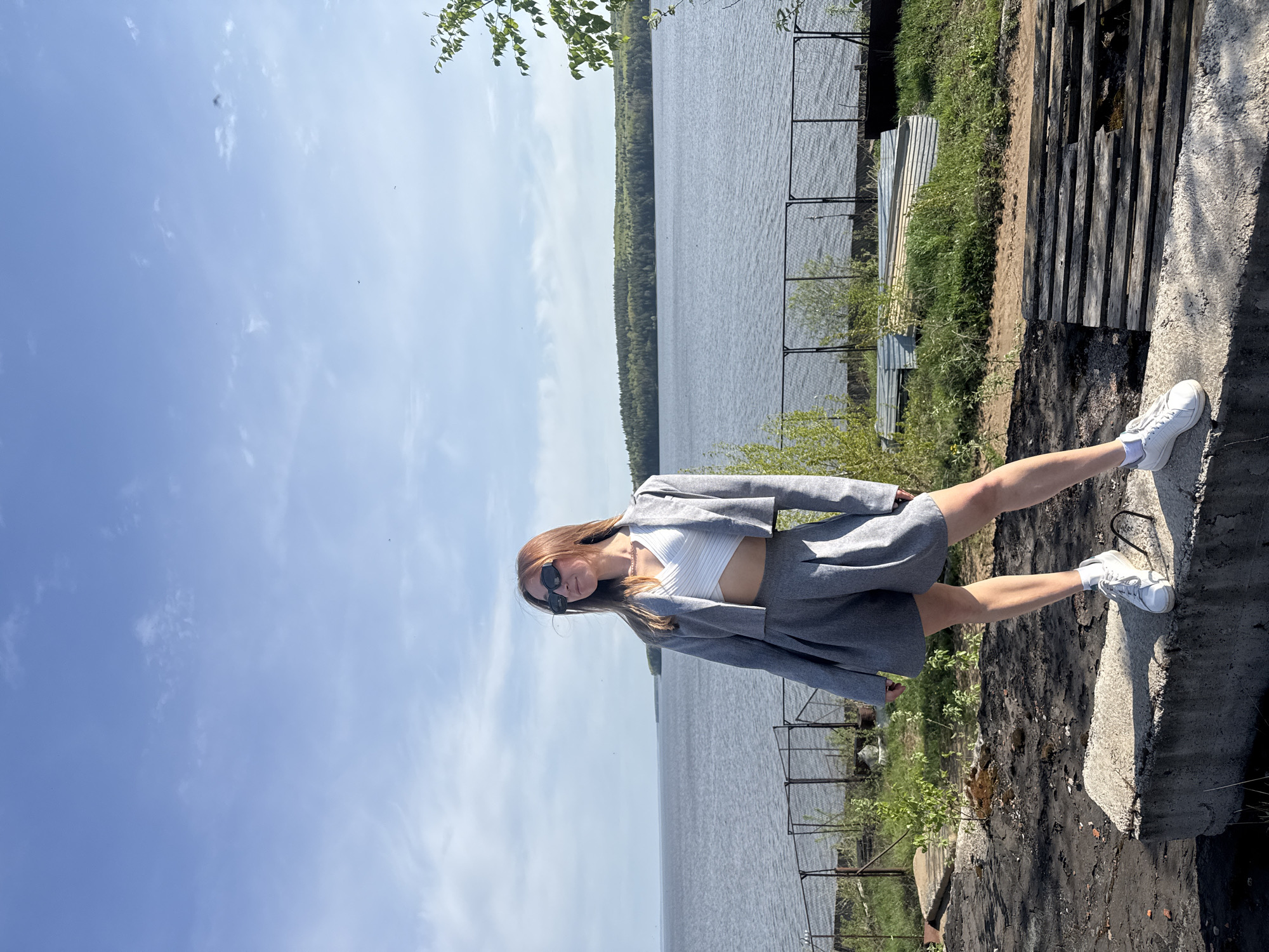 A young woman stands on a concrete structure near a body of water. She is wearing a white top, a gray jacket, a gray skirt, white socks, and white sneakers, and sunglasses. In the background, there is a lake and trees. A fence can be seen along the bank. There is also a wooden structure on the right. The sky is mostly clear. The image is rotated. Based on visual clues, this could be in Chaikovsky, Russia, as the prompt suggests.