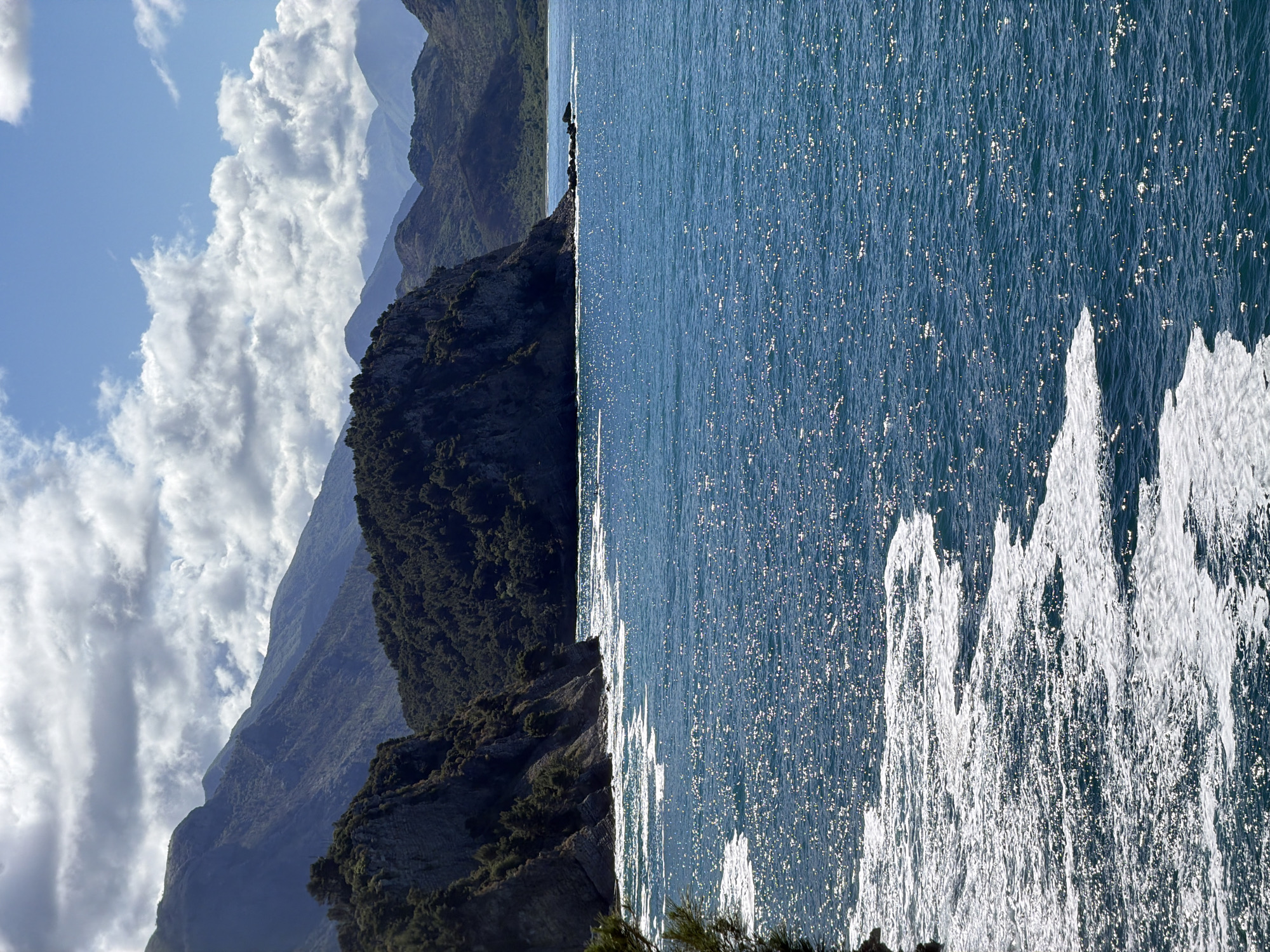 The image shows a view from Petrovac centar, Montenegro. On the left, mountains with dense green vegetation are visible, partially obscured by clouds. On the right, the blue sea sparkles under sunlight, showing the white spray of waves crashing against the rocks. The photo is oriented vertically.