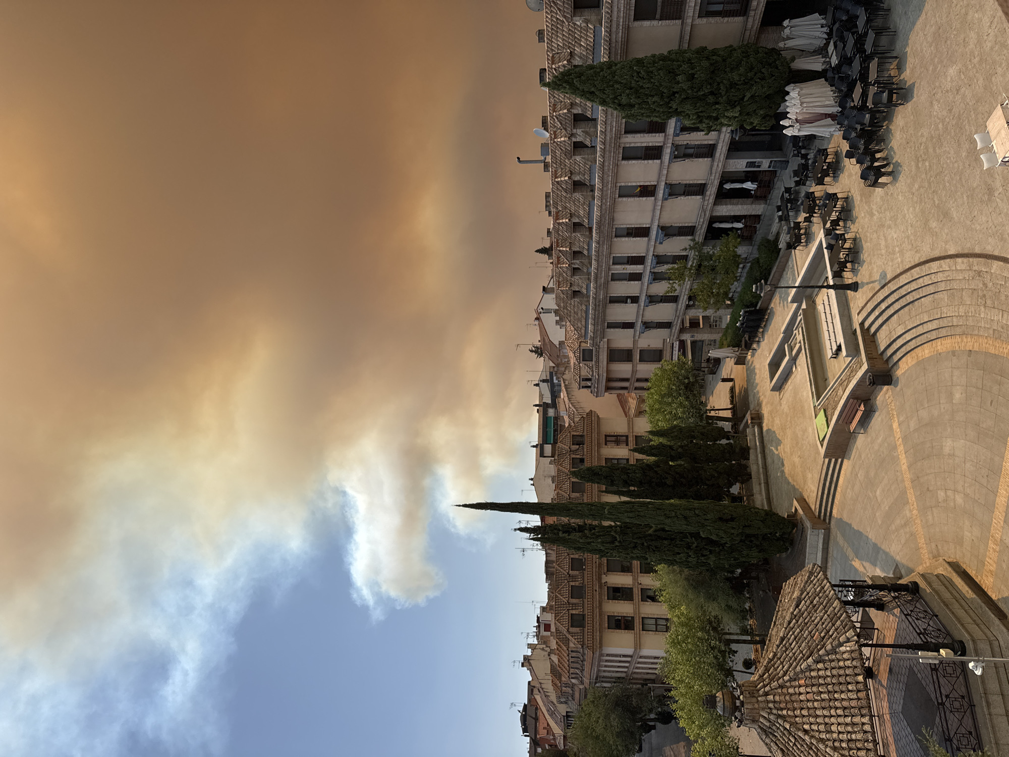 The image captures a panoramic view of a European city square, likely Las Rozas de Madrid, Spain, under a sky choked with thick, orange-brown smoke. The smoke, originating from a distant wildfire, casts an eerie, apocalyptic glow over the scene. In the foreground, a tiled gazebo with an ornate metal railing sits to the left, its dark roof contrasting with the hazy atmosphere. The square itself is paved with light-colored stones and features a water fountain structure, empty benches, and several trash receptacles.
To the right of the square, an outdoor seating area of a restaurant is visible. Numerous dark chairs and tables are neatly stacked or arranged, with some tables still set with white tablecloths and umbrellas. A tall, slender cypress tree stands prominently near the restaurant.
Behind the square, multi-story buildings with tiled roofs and light-colored facades line the streets. The architecture suggests a European urban setting. Windows are visible on the buildings, some appearing dark, others with blinds drawn. The upper floors of the buildings feature balconies or extensions.
The sky dominates the upper portion of the image. A clear blue sky is visible on the far left, but it is rapidly overtaken by a dense plume of smoke, colored in shades of orange, yellow, and brown. The smoke appears to be a significant event, obscuring the sun and dramatically altering the natural light. No people are visible in the scene. The overall impression is one of an unnerving, atmospheric event impacting a peaceful urban environment.
