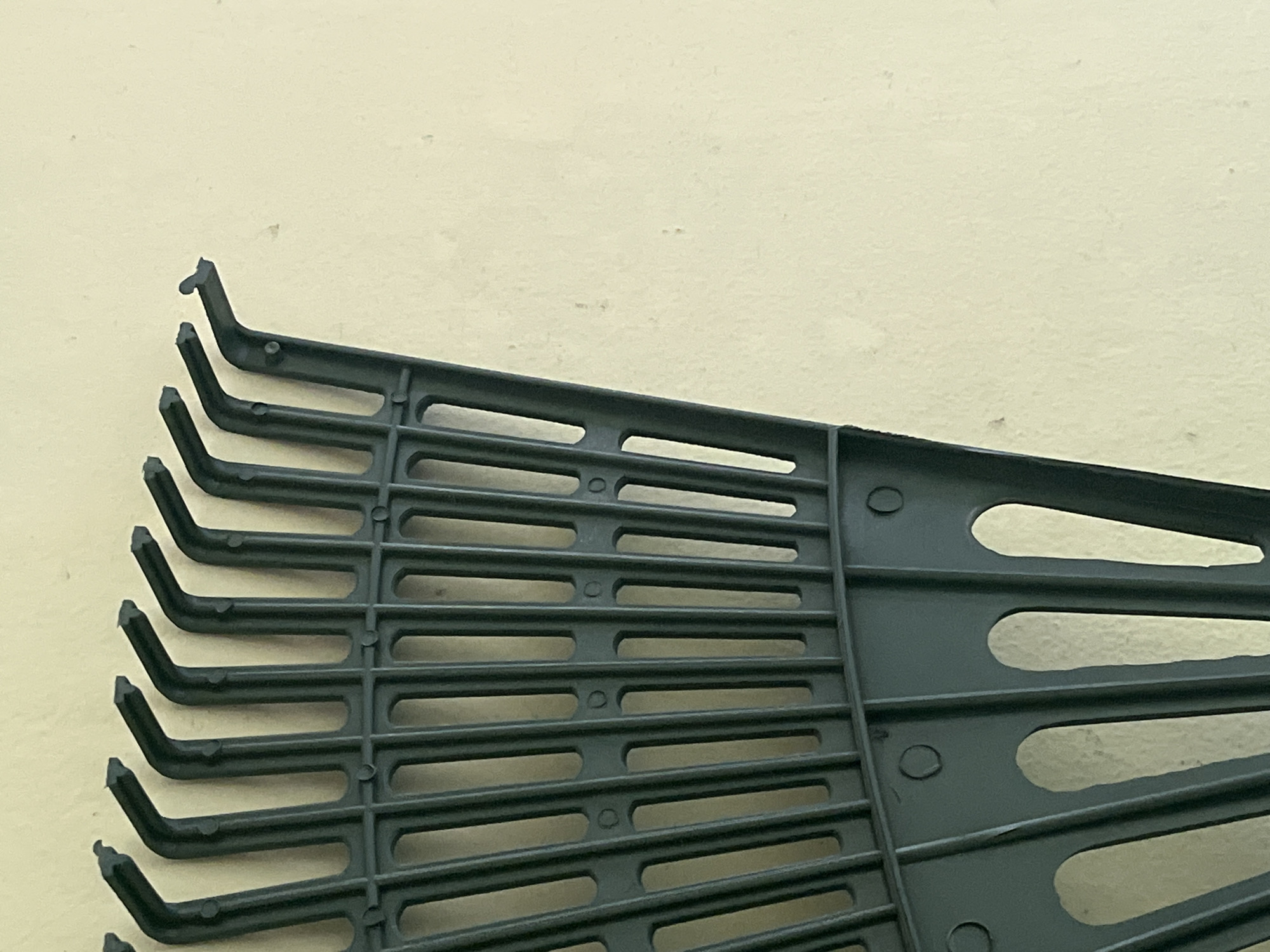 A close-up, high-angle shot shows the tines of a dark green plastic leaf rake leaning against a pale yellow, textured wall. The rake is positioned on the left side of the frame, with its tines fanning out towards the right. The wall occupies the majority of the right side of the image. The scene is indoors, likely in a residential or storage area, and does not depict any specific urban or natural landmarks. Therefore, the location in Teluk Air, Indonesia, cannot be determined from the visual information provided.