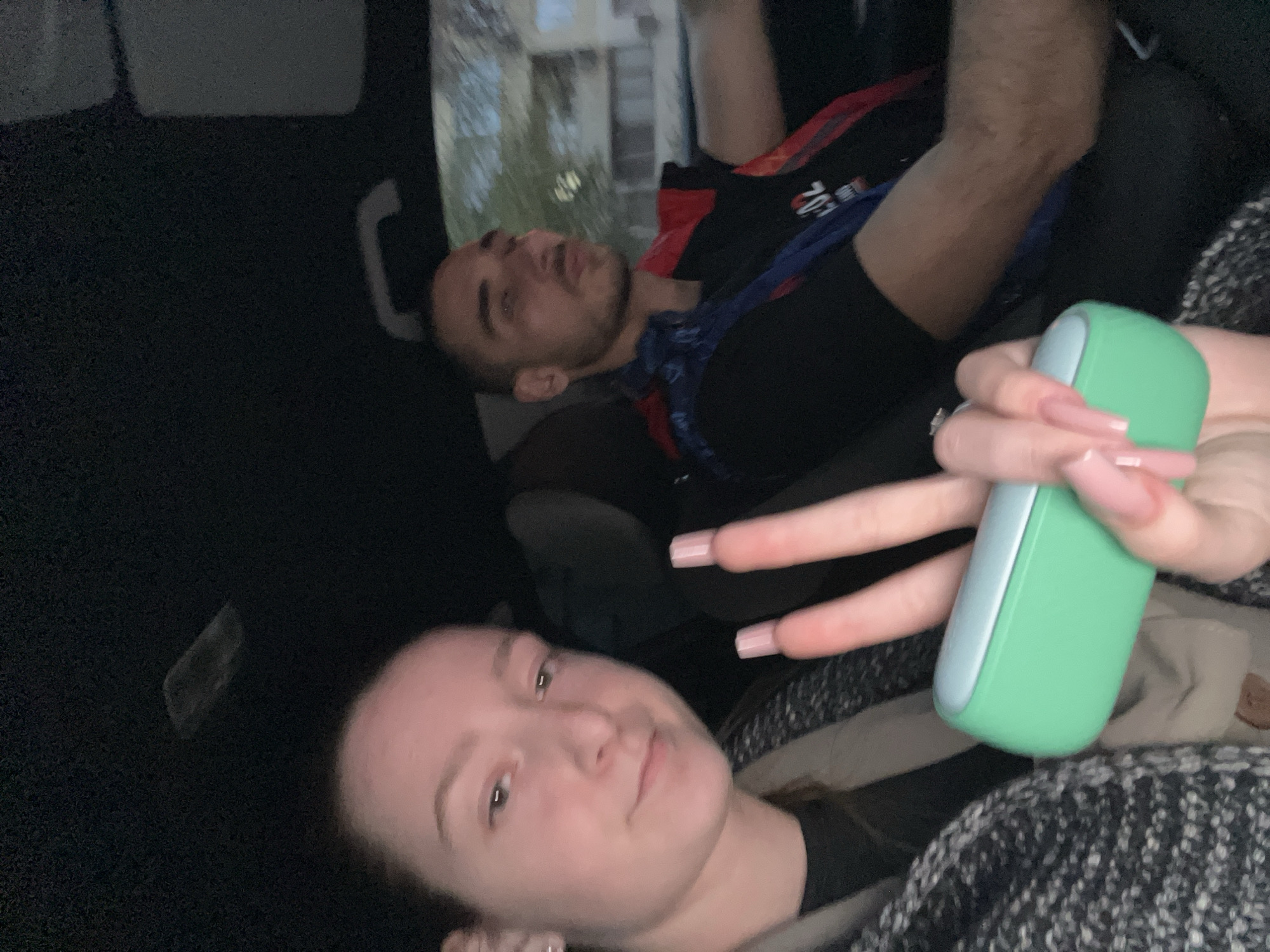 The image appears to be taken inside a car. There are two individuals visible. The woman is sitting in the back seat, facing the camera, and holding a green rectangular device in her hand, making a V-sign with her fingers. She has fair skin, light brown hair, and is wearing what seems to be a dark shirt. Another person, presumed to be a male, is lying down in the backseat, away from the camera. He's wearing a black shirt with some red and white design and a blue bandana. The environment is dimly lit, with the car interior being mostly black. There is a glimpse of a building or trees visible through the car window. The location is undetermined.