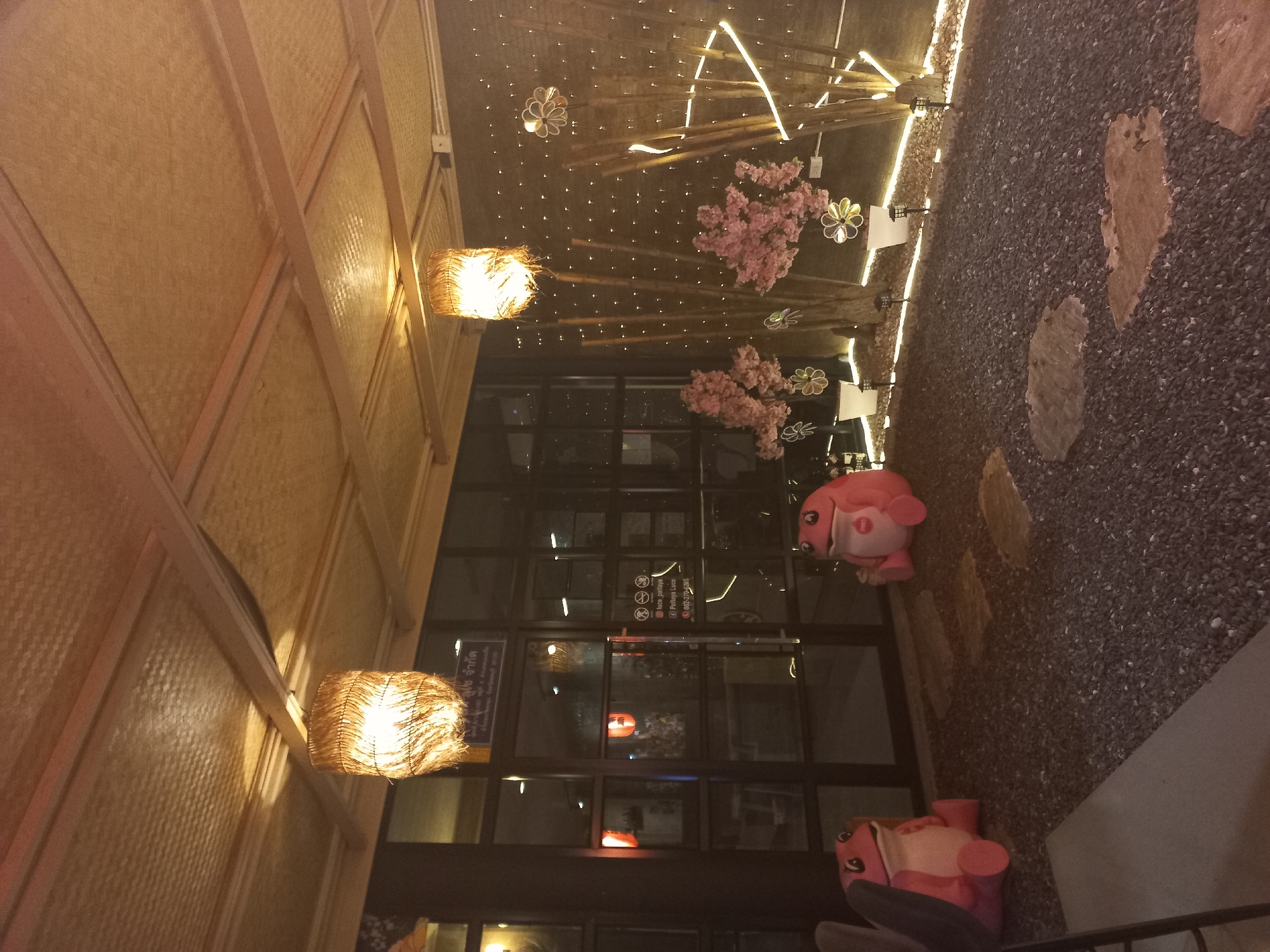 The image shows an outdoor area that is decorated with various lighting elements and props. Above, a woven bamboo ceiling supports decorative elements such as string lights, artificial cherry blossom branches, and flower-shaped light fixtures. Woven lamps hang from the ceiling. A glass door leads into the building. Next to the door, the exterior features a gravel surface, with large stepping stones. Pink cartoonish animal figures stand on either side of the doorway. The location of this image is in Pattaya City, Thailand as is indicated on the door.