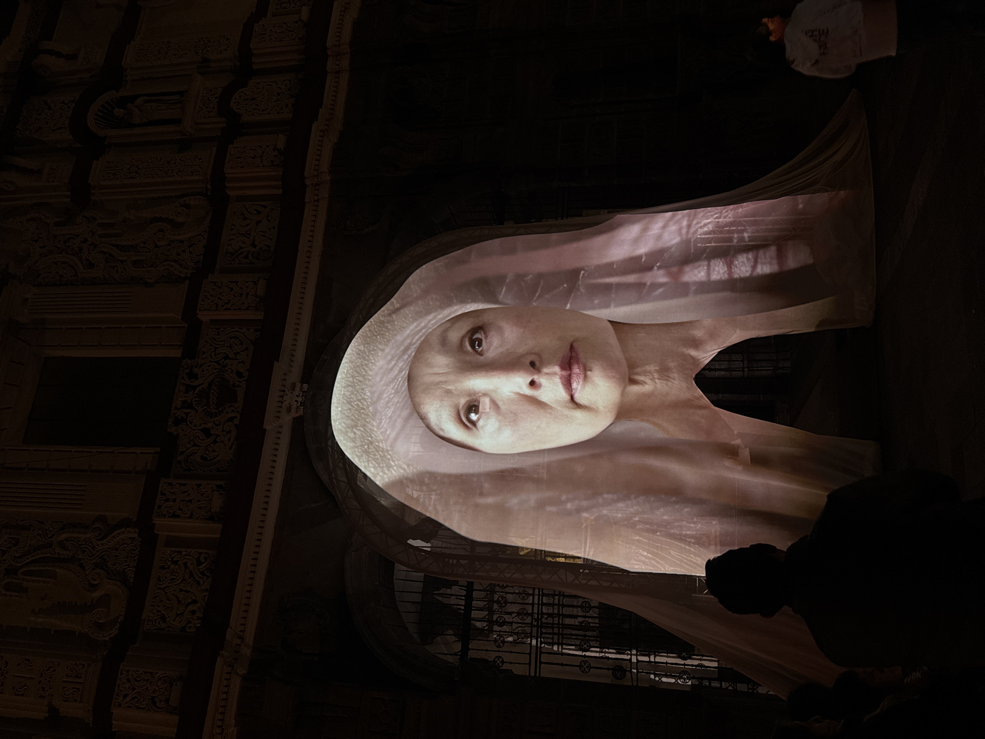 The image shows a large-scale projection of a woman's face wearing a light-colored hood or headdress, likely part of an art installation or performance. The projection is cast onto a structure with arched openings and intricate architectural details, suggesting a historical building. Based on the details of the stonework and architecture, it is likely located in Puebla, Mexico. The light from the projection illuminates the immediate area, while the surroundings remain dark. Silhouetted figures of onlookers can be seen in the foreground and background, indicating that this is a public display. The woman's face is the focal point, with a slightly melancholic or serene expression.