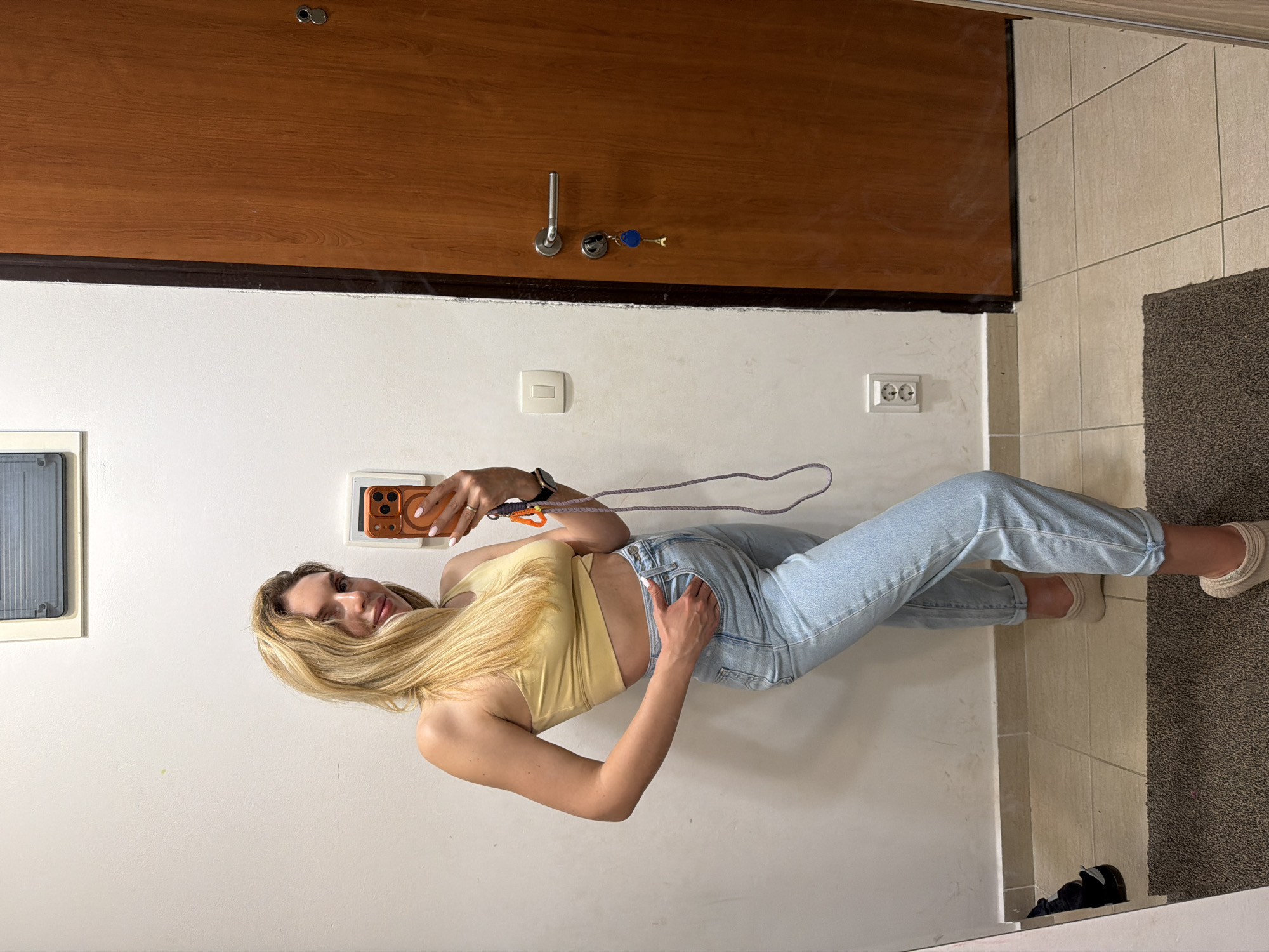 A blonde woman is taking a selfie indoors. She is wearing a yellow crop top and light blue jeans. She is standing in a hallway with a wooden door behind her and a gray doormat on the floor. Electrical outlets and a light switch are visible on the wall. The location is likely within a building in Budva, Montenegro.