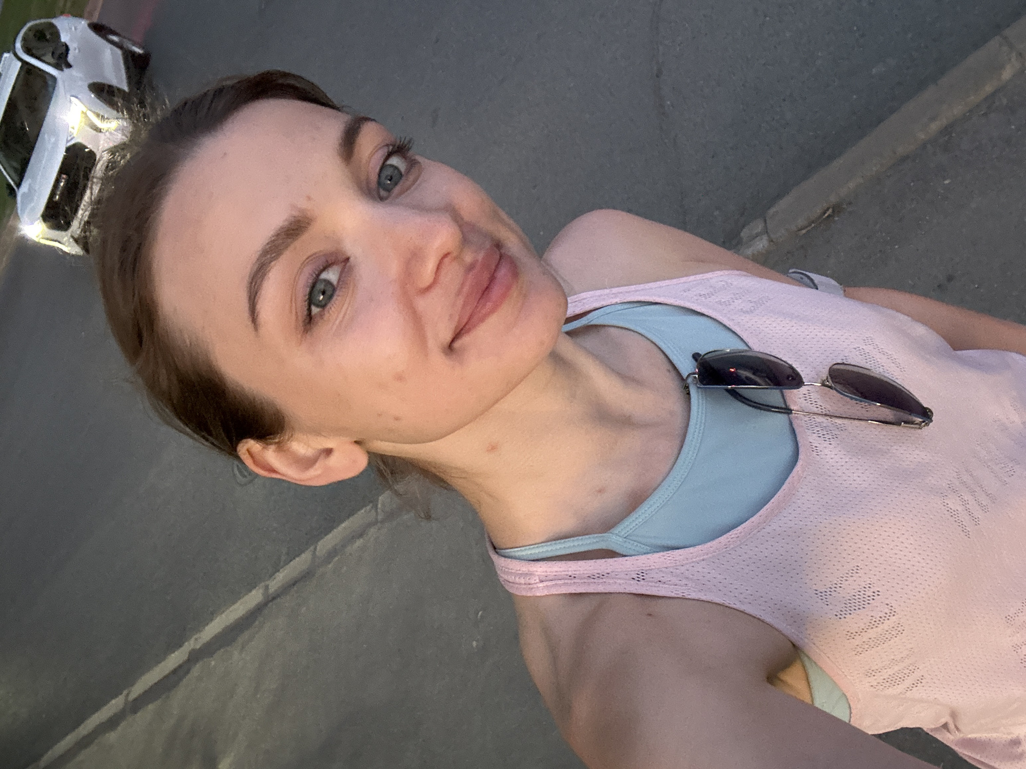 A woman is taking a selfie. She has fair skin, light-colored eyes, and her hair is pulled back. She's wearing a light pink tank top over a light blue sports bra, and sunglasses are resting on her chest. The background shows a gray asphalt surface, likely a street or sidewalk, with a white car parked nearby. The location cannot be determined.