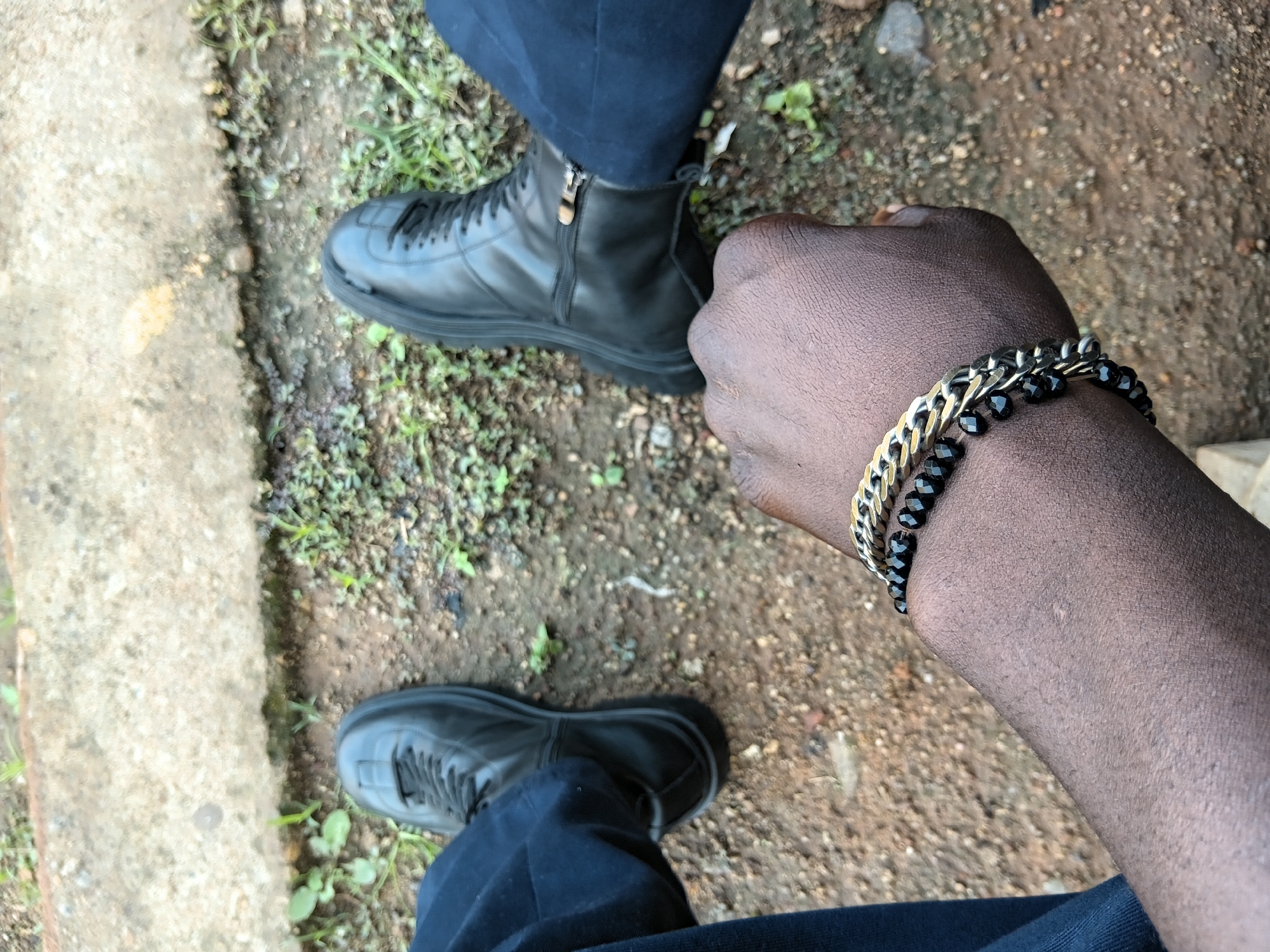 In this top-down shot of two feet on the ground, the left foot is wearing a black lace-up boot and dark blue pants. The right foot is wearing a black zip-up boot, also with dark blue pants. On the right arm, a hand with two bracelets is visible. The bracelets consist of a gold chain-link band and a black beaded band. The ground beneath the feet is composed of dirt, small stones, and sparse green vegetation. A concrete edge is visible in the background.