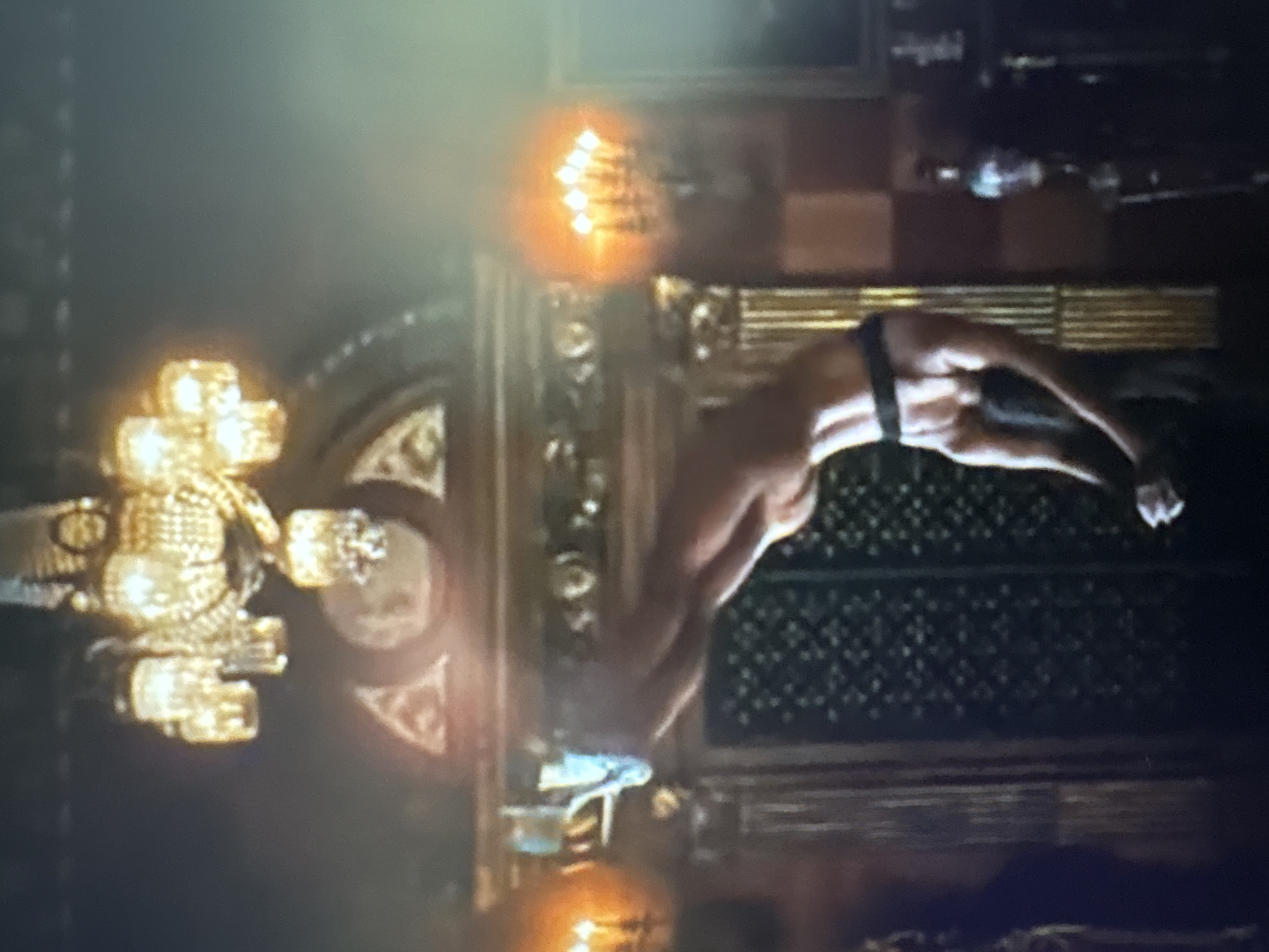 In a dimly lit, opulent room, a person is performing a backflip or acrobatic move. They are suspended mid-air, with their body arched backward and their legs extended. The person is wearing dark bottoms. The room features a grand chandelier with multiple lights hanging from the ceiling. On the walls are ornate sconces with lit candles, casting a warm glow. There are also tall, decorative columns and a patterned screen or wall covering. The overall impression is one of a luxurious interior. The image does not contain any information that would allow for identification of the location in the city of Sofia, Bulgaria.