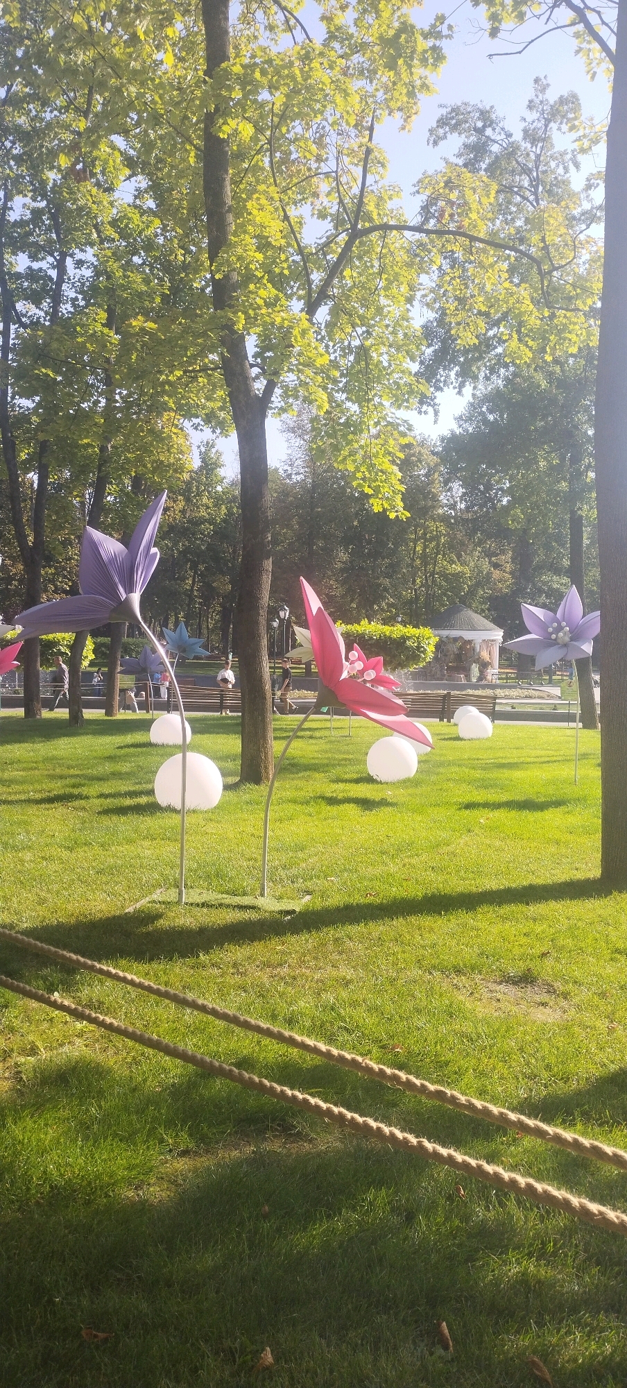 The image captures a park scene with large, decorative flower sculptures on a sunny day. The sculptures, appearing to be made of fabric, are in vibrant pink and purple hues and are positioned on thin metal poles, some adorned with white spherical lights at their bases. The ground is covered in a lush green lawn, with shadows cast by the trees and sculptures. In the background, people can be seen walking and sitting on benches, with a small gazebo or pavilion visible further back. The overall environment suggests a recreational area within a city, with mature trees providing shade and a clear sky overhead. The location is identified as Kharkiv, Ukraine, implying this is likely a public park or garden.