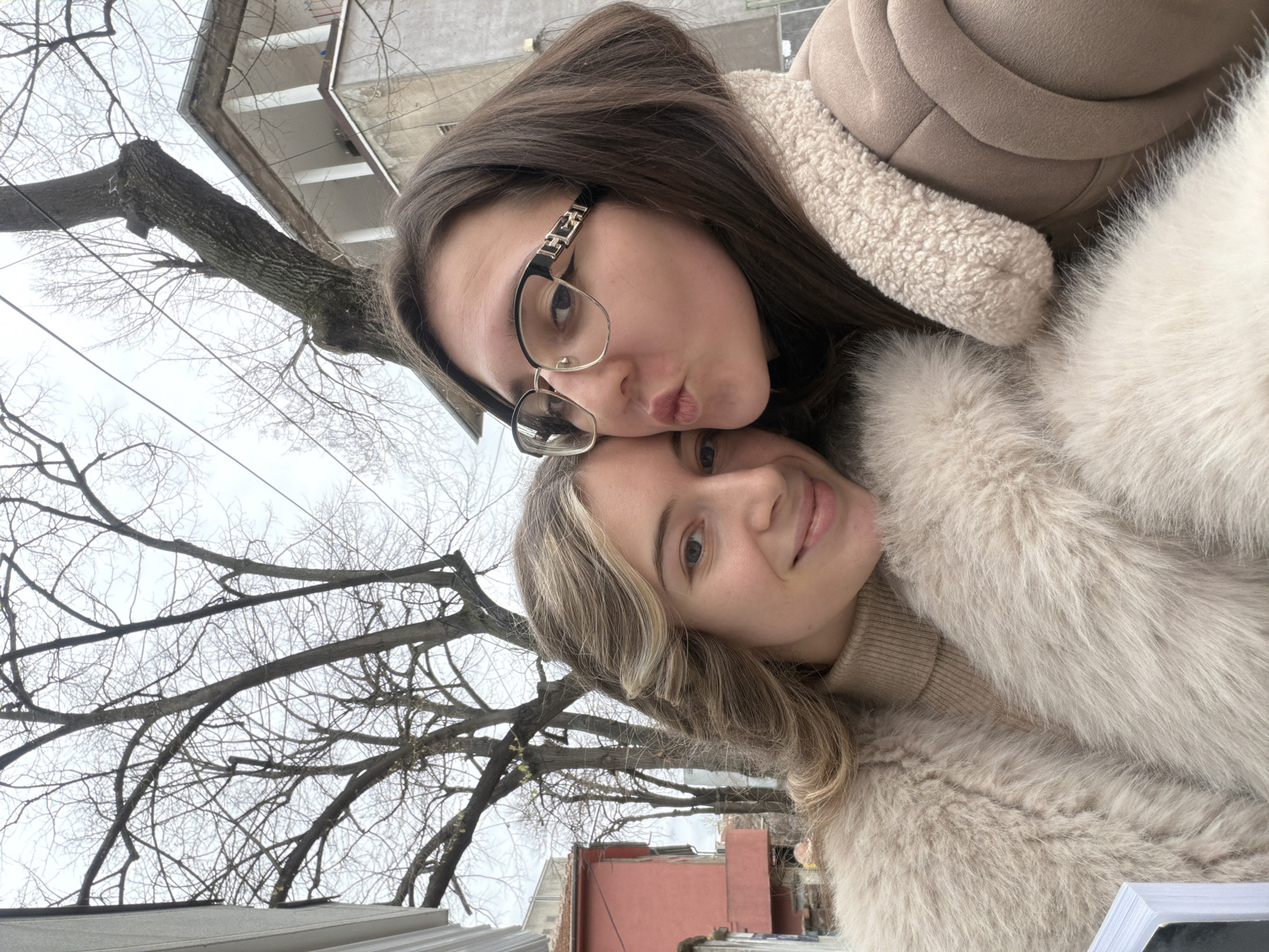 Two women are posing closely together outdoors, likely in a city setting. One woman has long brown hair and is wearing glasses; she is pouting her lips as if to kiss. The other woman has blonde hair and is smiling slightly. Both women are wearing warm, fur-like coats. Behind them is a bare tree and some buildings. Based on the architectural style visible, the location appears to be Plovdiv, Bulgaria.