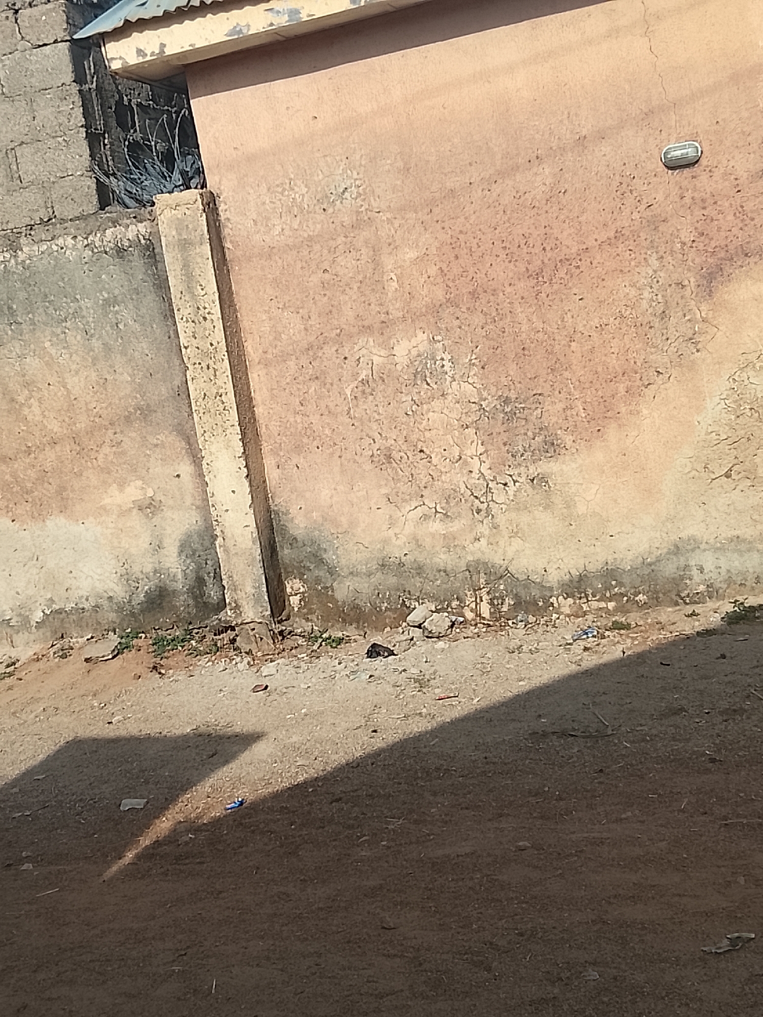 The image captures a static outdoor scene featuring a weathered building wall and unpaved ground under strong sunlight. The prominent wall, appearing faded peach or light brown, shows significant signs of age, including numerous cracks, peeling paint, and dark, damp-stained discoloration along its lower section. A tall, slender concrete pillar or column stands vertically, integrated with this main wall, also showing signs of wear.
To the left, a portion of another wall constructed from grey concrete blocks is visible. Between the main wall and the concrete block wall, there's a gap revealing a tangle of what appears to be barbed wire or sharp metal wires. A small section of a corrugated metal roof overhangs this opening from the top. Mounted on the upper right side of the main wall is a small, oval-shaped outdoor light fixture.
The foreground consists of rough, dusty ground, sparsely covered with small rocks and patches of green vegetation. Scattered debris is visible on the ground, including a distinct small blue object, possibly a pen or a piece of plastic, near the center. Deep, sharp shadows are cast across the ground, indicating a bright, sunny day, likely during the morning or late afternoon when the sun is at an angle. No human activity or visible text is present in the scene. The overall appearance of the structures and environment is consistent with general architecture and conditions found in locations like Bauchi, Nigeria.