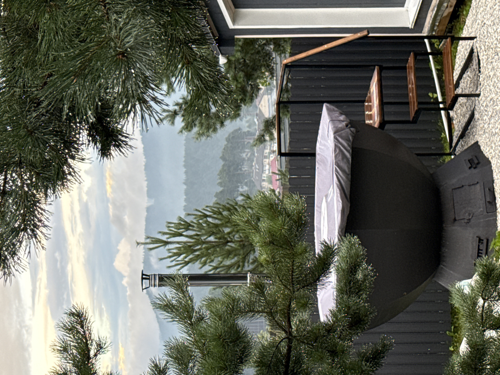 This is a vertical, eye-level, outdoor shot of a balcony in Kusa, Russia. The foreground is dominated by the needles of pine trees, some of which are laden with water droplets, suggesting recent rain or mist. Beyond the trees, a dark, modern balcony railing encloses a space where a large, dark, rounded object, possibly a covered hot tub or an outdoor grill, is partially shrouded in a light-colored fabric cover. To the right of this object, a simple wooden bench with a metal frame is visible.
The background opens up to a hazy, green landscape of rolling hills and a small cluster of houses, hinting at a village or town nestled in the valley. The sky is overcast with soft, diffused light, indicating either early morning or late afternoon, with a touch of yellow in the clouds suggesting the sun may be trying to break through. The weather appears damp and cool.
The overall mood is one of quiet observation, with the natural elements of the pine trees and the distant landscape contrasting with the man-made structures of the balcony and the covered object. There is no visible text in the image.