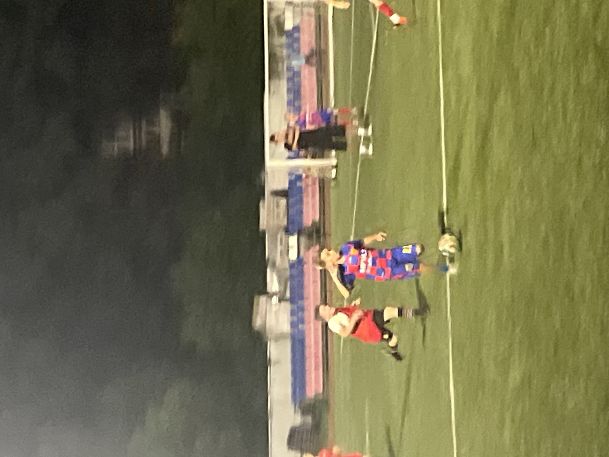 This is a vertical, low-light, medium-grain photograph taken at night or dusk, possibly in Krasnodar, Russia. The scene depicts a sports field, likely a soccer pitch, with artificial turf. In the foreground, two individuals are visible, appearing to be athletes. The one closer to the camera is wearing a red jersey and black shorts, with their left leg extended as if kicking a ball, which is out of frame or obscured. The other athlete, further back and slightly to the right, is wearing a blue and red patterned jersey and black shorts. They are also in motion, with their right leg bent and their arms positioned as if running or jumping.
Behind the athletes, a structure resembling a stadium bench or viewing area is visible, with individuals in dark clothing seated or standing. The background is dark, with faint shapes suggesting trees or buildings, further indicating the low-light conditions. A white rope or line runs diagonally across the field. The overall impression is of a soccer match or practice in progress, captured during an evening or nighttime event. No visible text is present in the image.