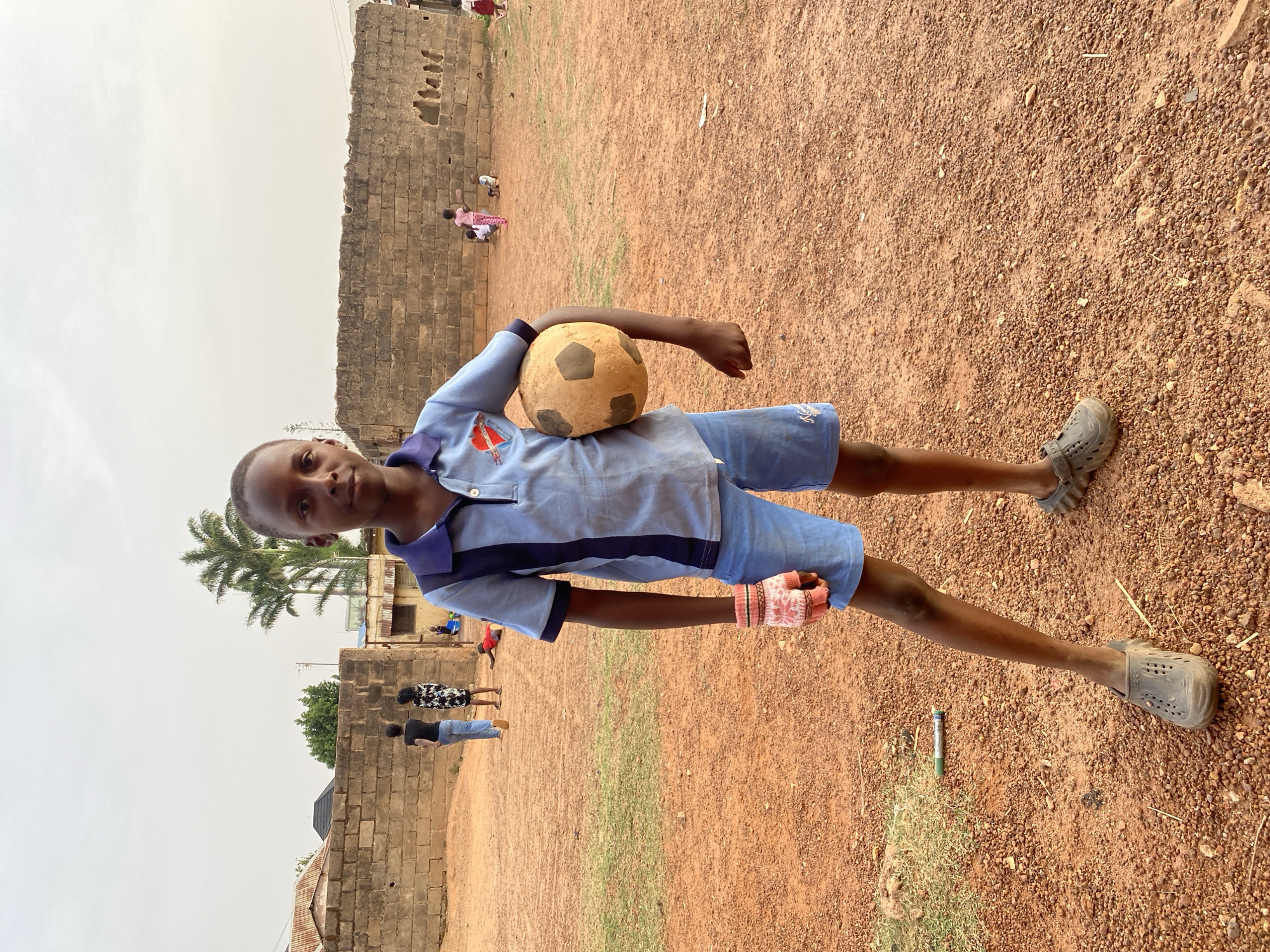 A young boy stands centrally in an outdoor setting, holding a worn, light-colored football with dark patches under his right arm. He looks directly at the camera with a neutral expression. He is dressed in a light blue collared shirt with dark blue cuffs and collar, light blue shorts, and grey clogs. A distinctive red and white patterned fingerless glove is visible on his left hand. A small, indistinct emblem is present on the left side of his shirt.
The boy is positioned on an unpaved, reddish-brown dirt and gravel ground. In the background, a low, light brown stone wall runs horizontally across the scene. Beyond the wall, several other people, including children and adults, are visible moving or standing. Some simple buildings and green foliage, including palm trees, can be seen further in the distance. The sky is overcast, providing soft, diffused lighting, indicating daytime. A green pen or marker lies on the ground near the boy's right foot. The scene suggests a casual outdoor environment, possibly a community area or a schoolyard in Bukuru, Nigeria.