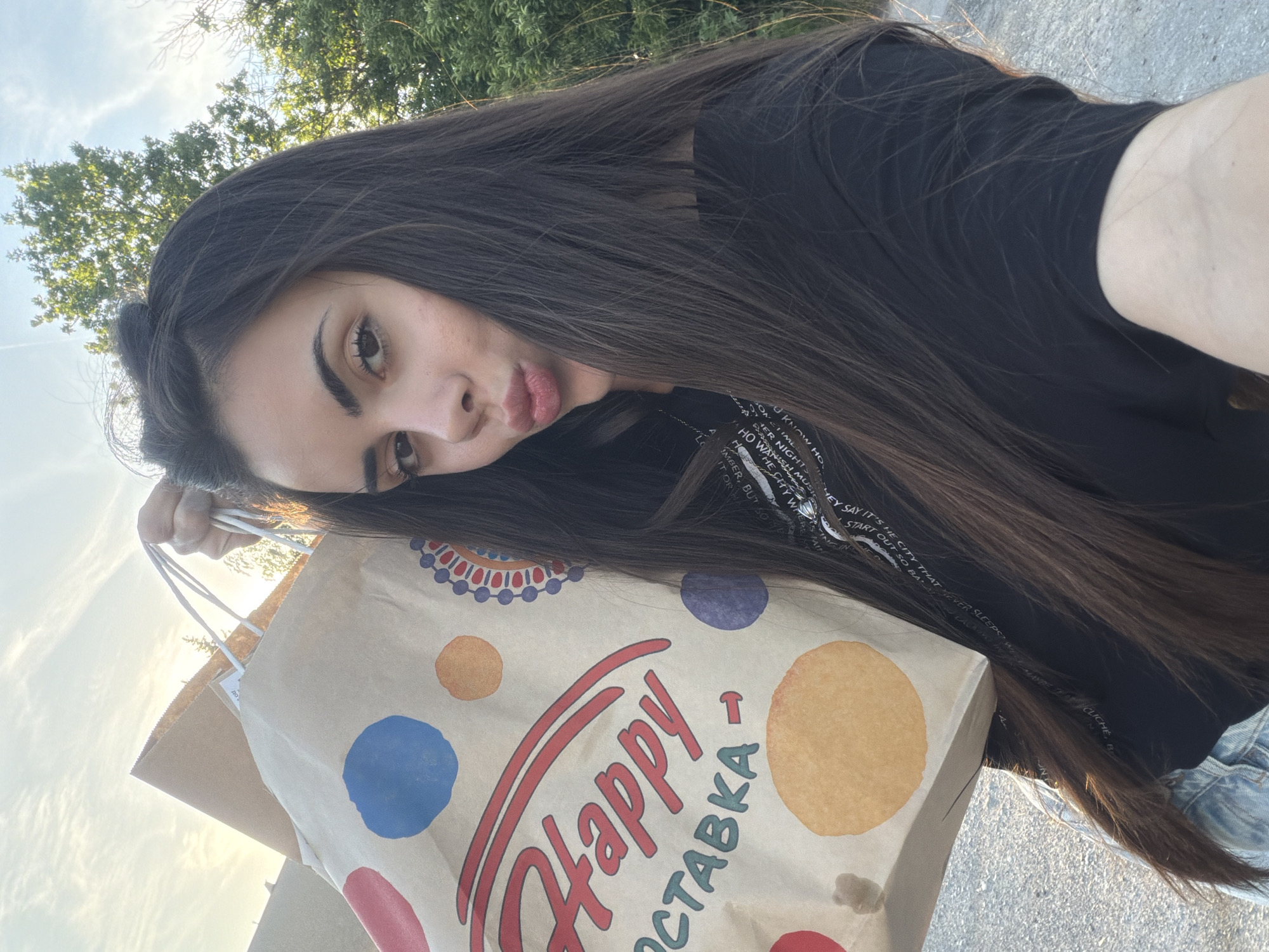 A young woman with long, dark brown hair is holding a brown paper bag with the word "Happy" and a Cyrillic word "OCTABKA" printed on it. The bag also has colorful polka dots. She's making a kissing face towards the camera. She's wearing a black T-shirt with text written on it. The background includes a tree and a street. Based on the Cyrillic text on the bag and the person's appearance, it can be inferred that the photo was taken in Sofia, Bulgaria.