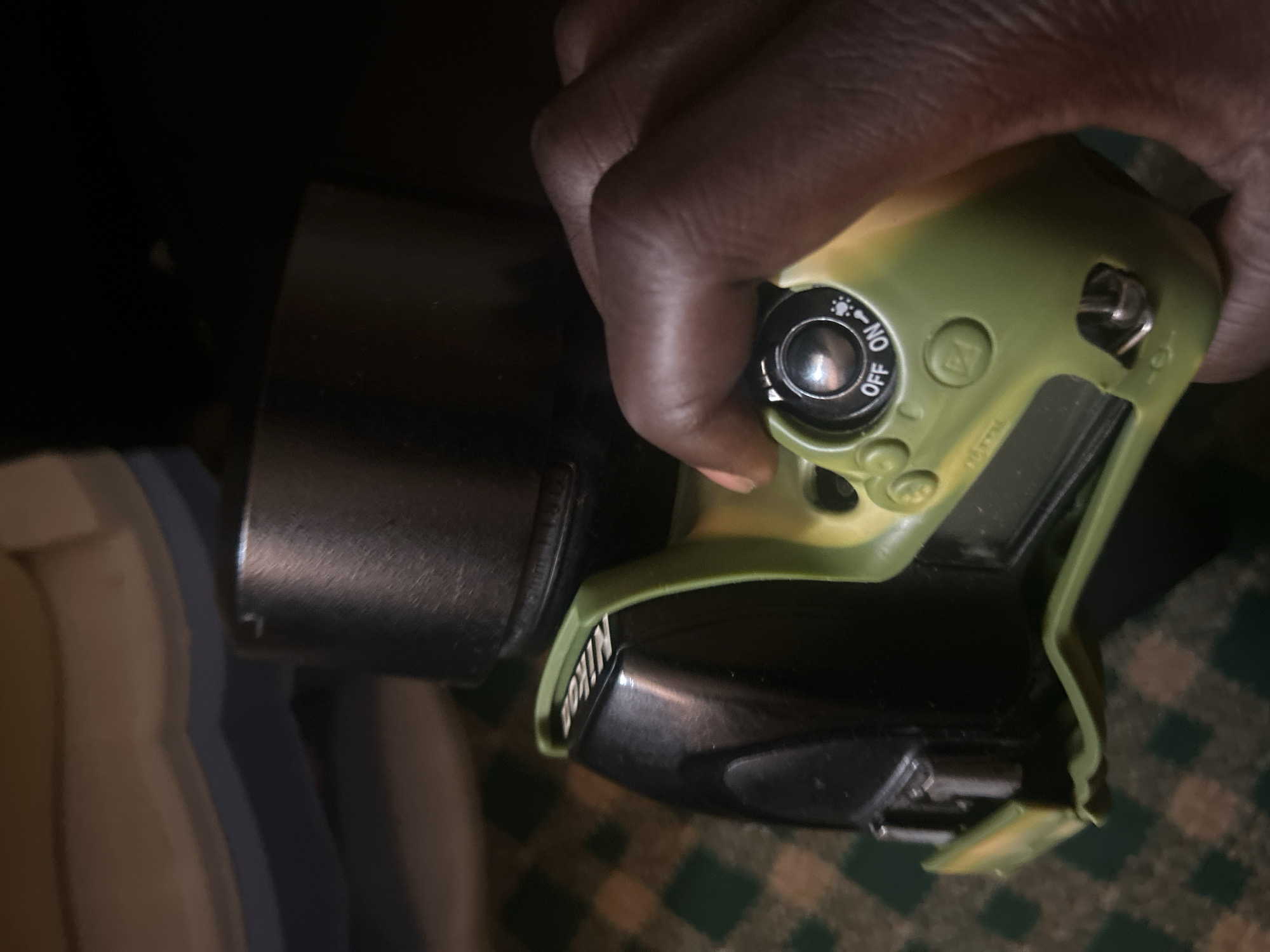 A person's hand with dark skin is shown from the wrist down, grasping the green silicone case of a black Nikon digital camera. The camera is positioned horizontally, with the lens pointing upwards and away from the viewer. The hand is operating a switch on the camera body that is labeled "OFF" and "ON" with a symbol indicating a flash. The camera itself is partially covered by the green case, which is slightly peeled back on the left side. The background consists of a green and white checkered surface, likely a tablecloth, and a blurred object with blue and tan stripes. The scene appears to be indoors. There is no discernible information about the location in Jos, Nigeria.