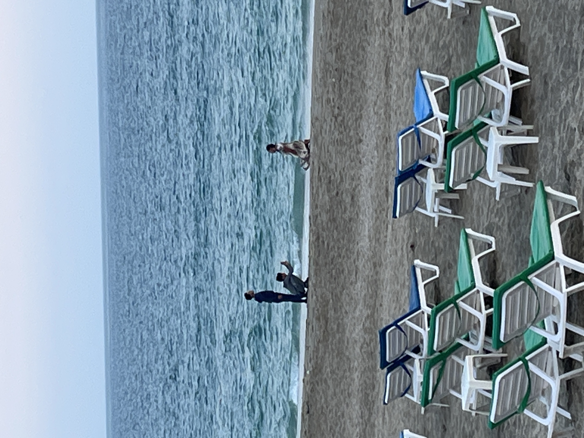 An outdoor scene depicts a beach in Alanya, Turkey, under what appears to be an overcast or hazy sky. The expansive blue-green sea, textured with gentle waves, occupies the left side of the frame, meeting a sandy beach on the right.
Along the sandy shore, primarily in the mid-ground and foreground, numerous plastic beach chairs are visible. These chairs are mostly white with green backrests, though some feature blue backrests. They are stacked and arranged in neat rows, indicating a prepared setup for beachgoers, likely part of a resort or public beach area.
Near the water's edge, three individuals are present. One person, dressed in light clothing, is positioned further down the beach, standing in the shallow water and seemingly observing the sea or the coastline. Closer to the viewer, two other individuals stand on the wet sand at the water's edge. One is dressed in dark clothing, and the other in lighter attire; they appear to be interacting with each other or looking out towards the sea.
The lighting is bright but diffused, suggesting an overcast day or a time when the sun is not directly overhead, lending a soft quality to the scene. There is no discernible text visible in the image.
