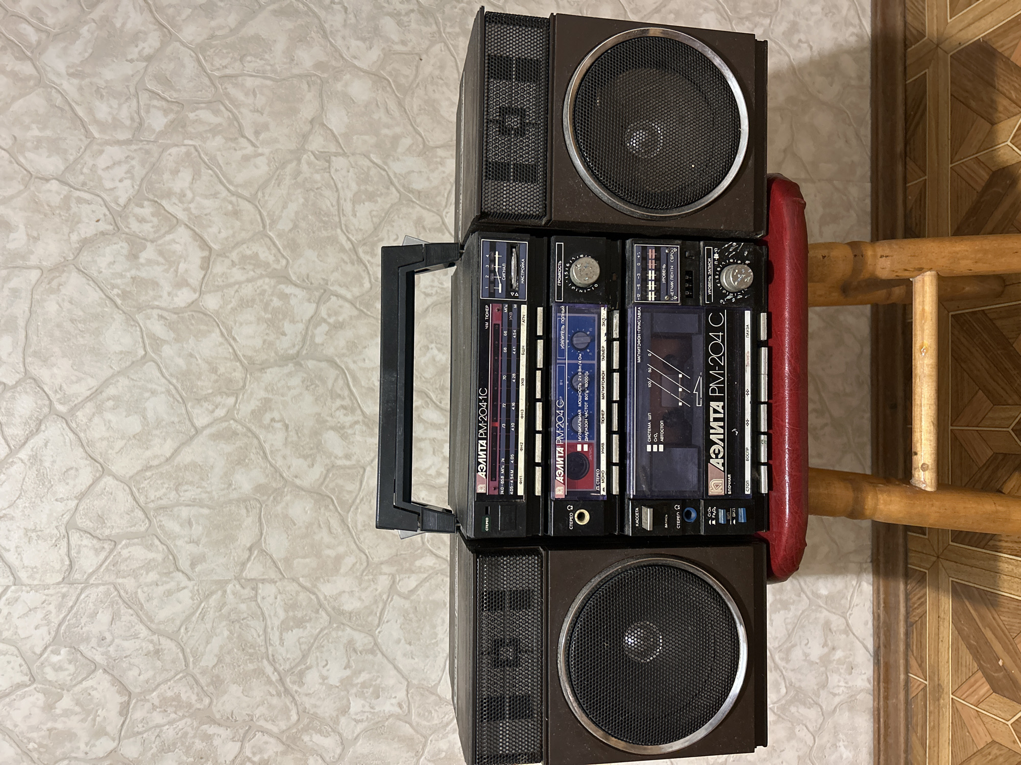 A vintage Soviet-era "Элита PM-204C" boombox, a portable cassette player and radio, is displayed prominently on a wooden stool. The boombox features two large speakers on either side of a central unit with controls for tuning, volume, and cassette playback. The unit also has a built-in radio tuner with frequency indicators in MHz and kHz, and a cassette deck with indicators for recording levels. The background is a textured wallpaper with a pattern resembling light-colored stones. The floor beneath the stool has a geometric tile pattern. The scene is indoors, likely in a residential setting, in the city of Lyubertsy, Russia. There are no people visible in the image.
